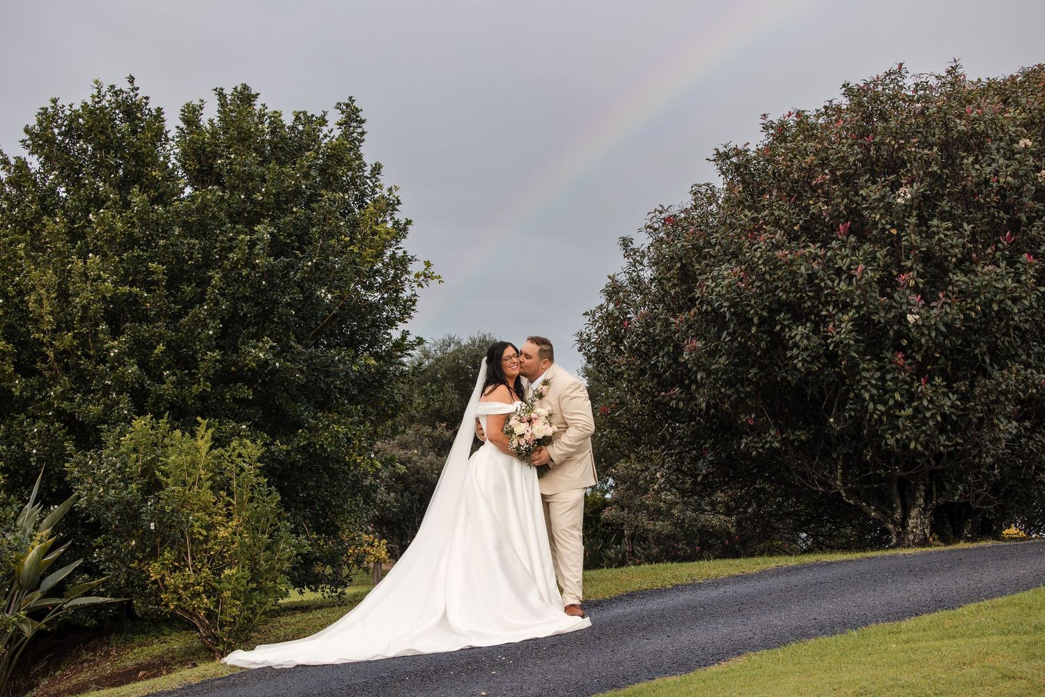 Bride and groom kissing on a garden path with a rainbow arching over lush green trees on their wedding day.