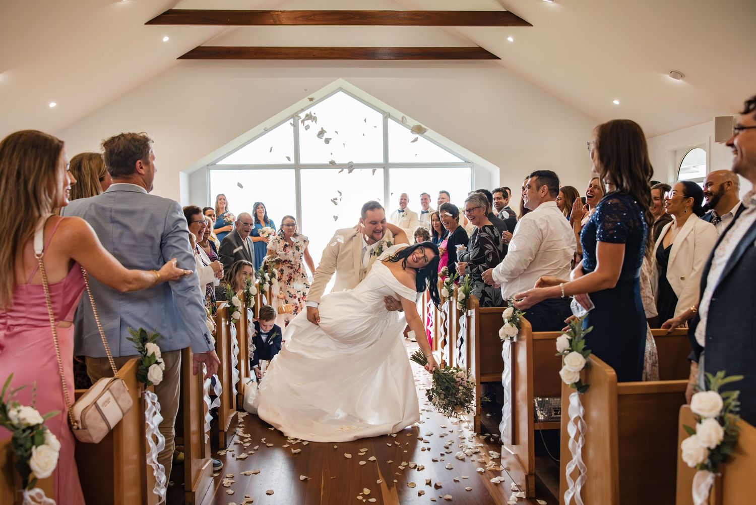 Bride and groom share a joyful moment in a white chapel aisle surrounded by guests tossing flower petals.