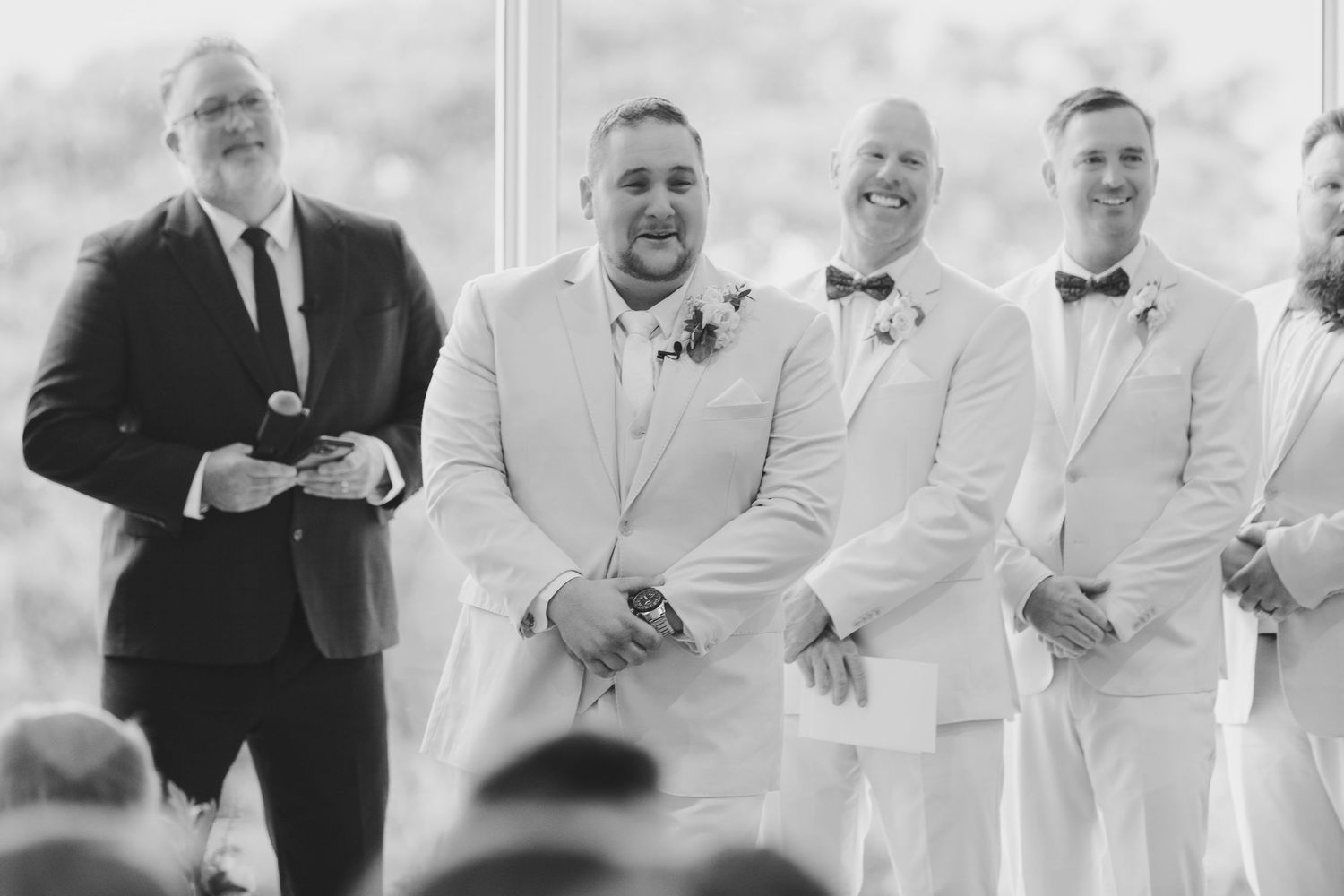 Groom and groomsmen in white suits and bow ties smiling at wedding ceremony altar in black and white photo.