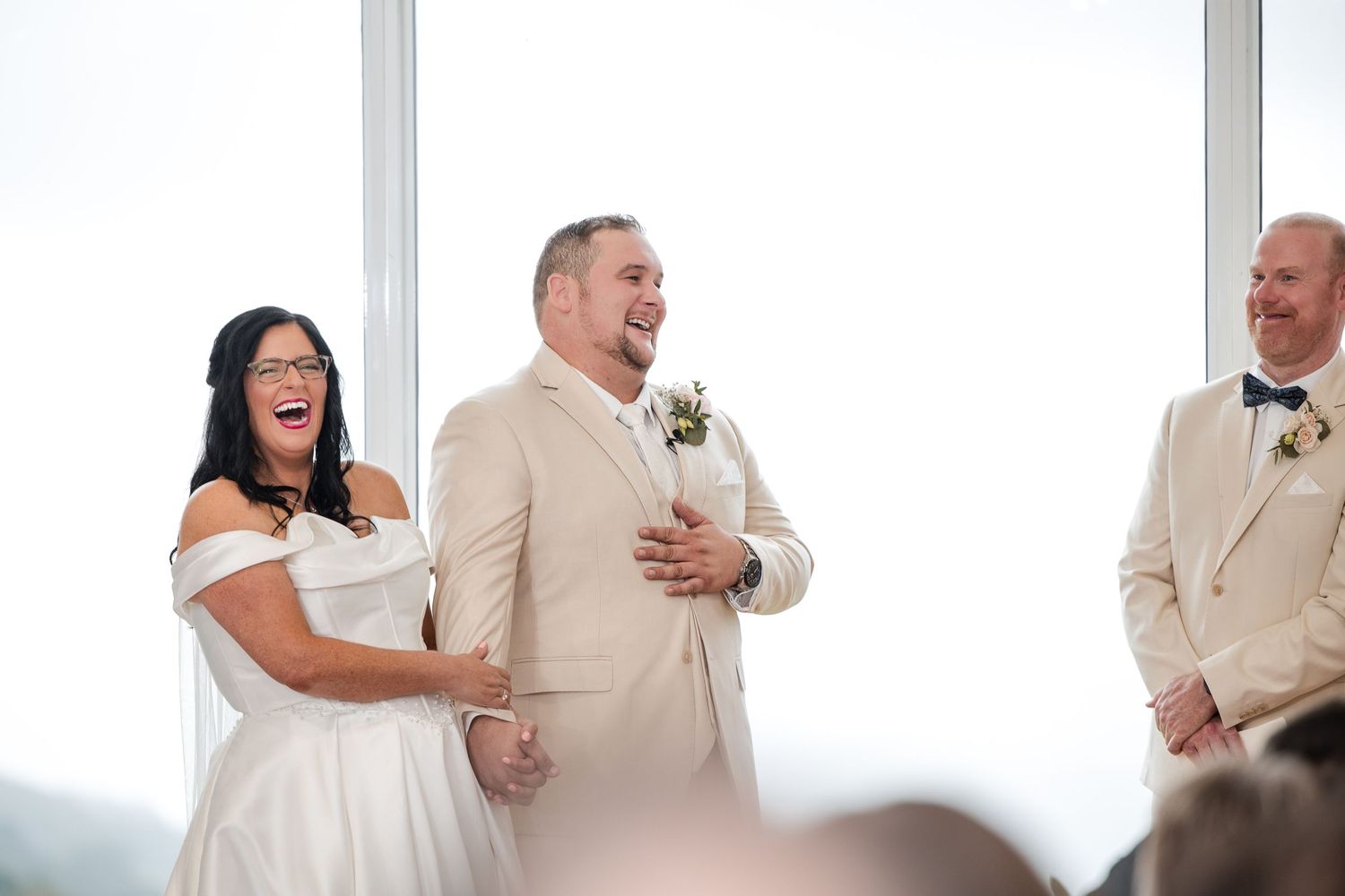Bride and groom laughing joyfully during wedding ceremony, bride in white off-shoulder gown, groom in beige suit with boutonniere.