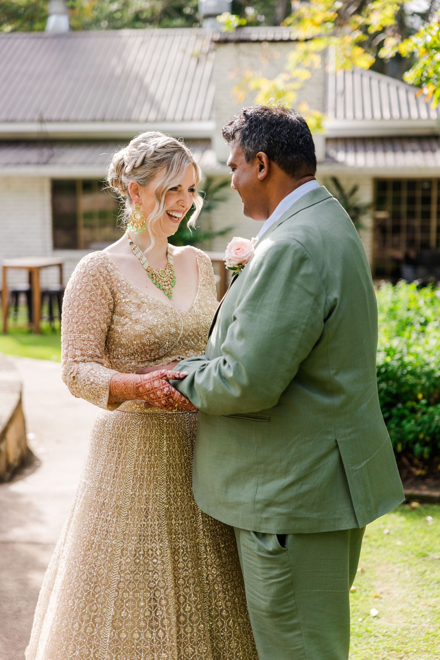 Bride in gold lehenga and groom in sage green suit hold hands and smile at each other outdoors on their wedding day.