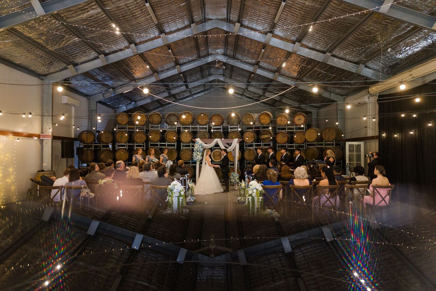 Bride and groom exchange vows in a stunning winery barrel room ceremony with guests seated in wooden chairs.