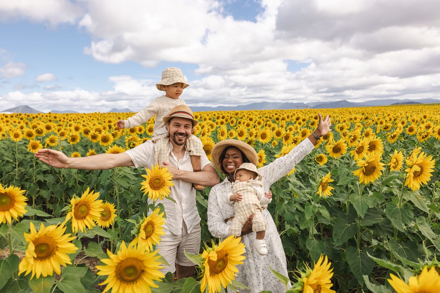 Family of four joyfully posing in a vast sunflower field under a partly cloudy sky, wearing matching white outfits.