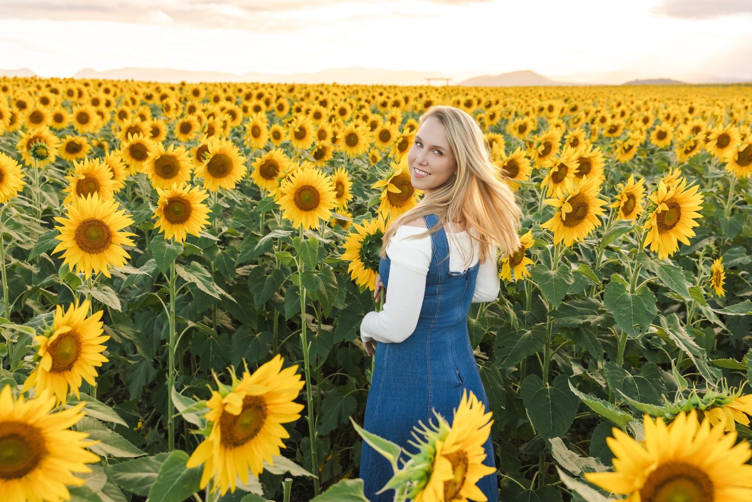 Blonde woman in denim overalls standing in a vast sunflower field at golden sunset.