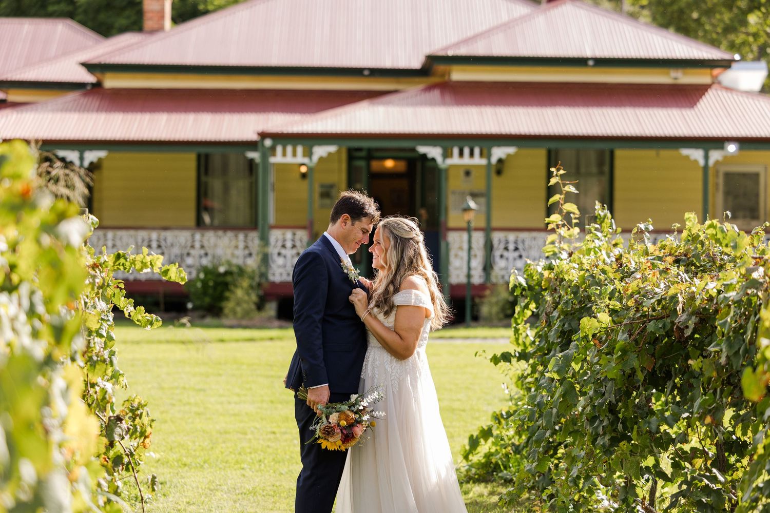 A bride and groom embrace in front of a historic building surrounded by lush greenery.