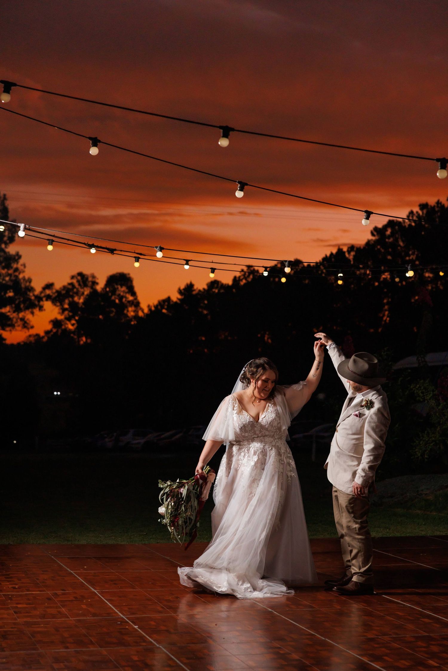 A couple dances under string lights against a dramatic sunset sky.