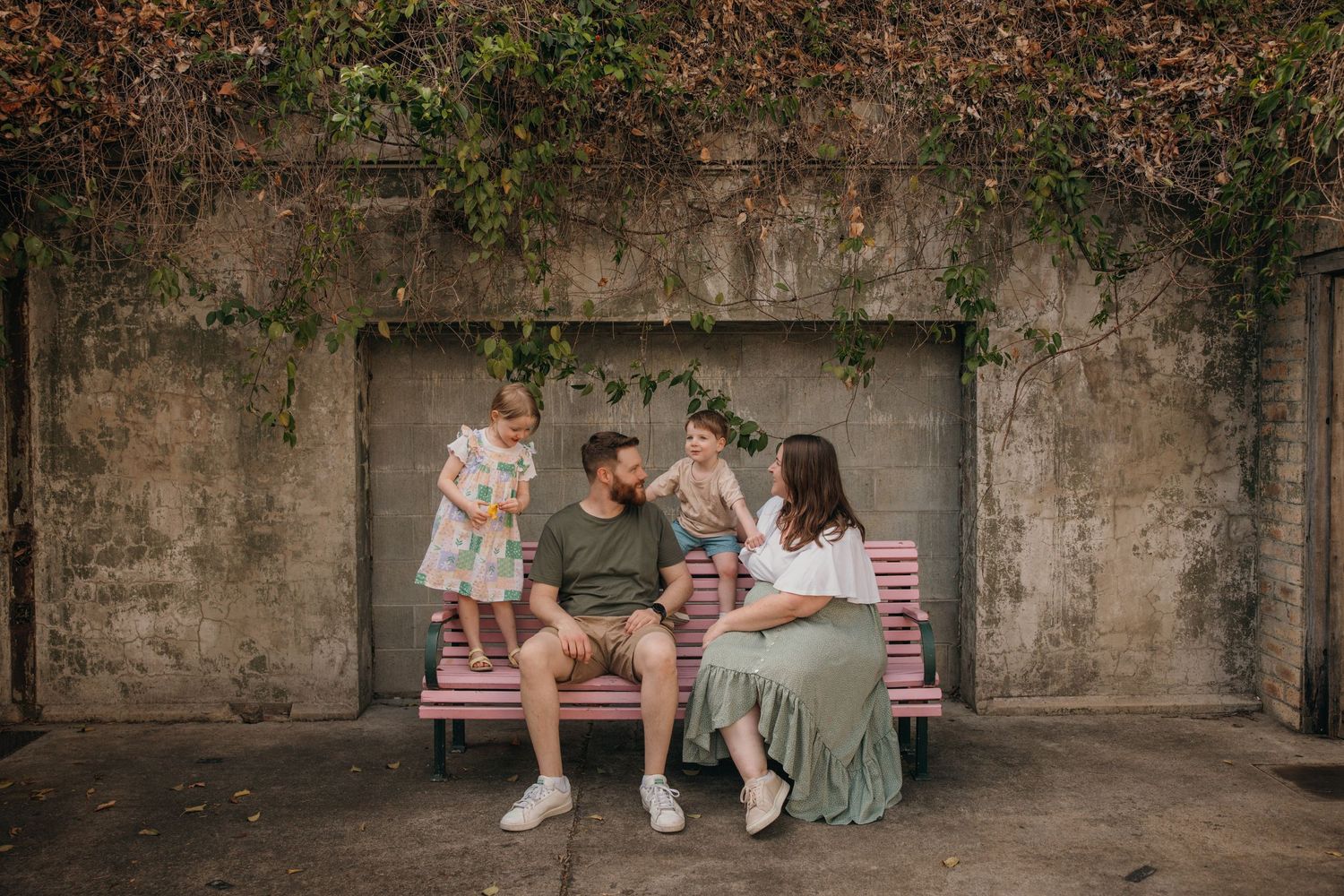 A family sitting together on a bench against a weathered stone wall with vines growing above.