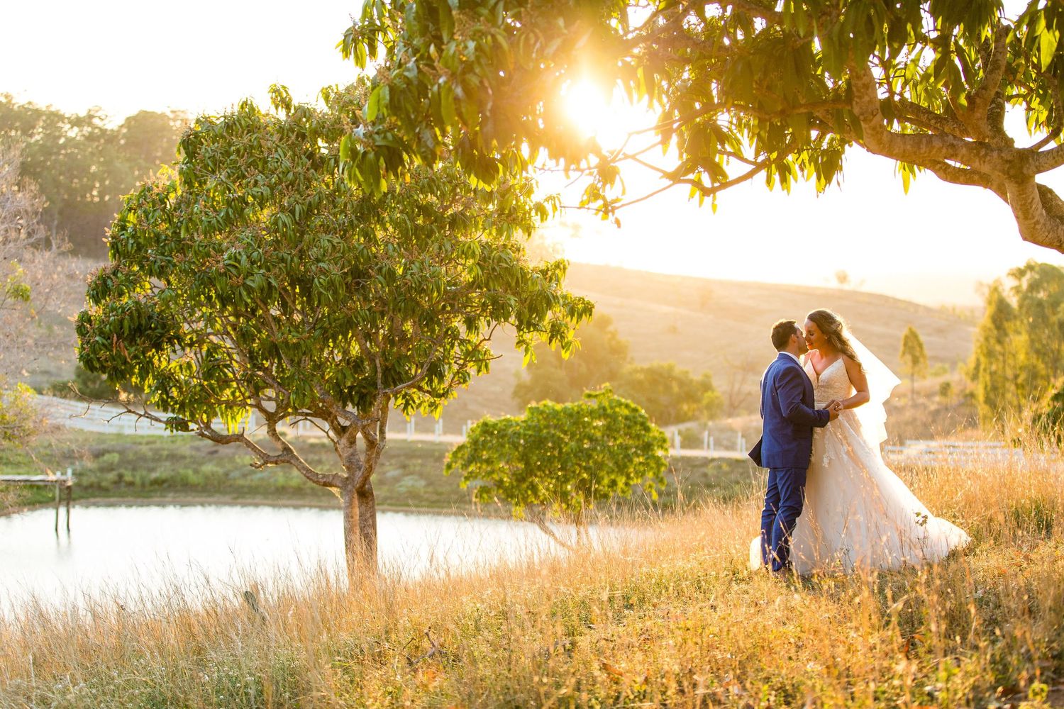 A romantic sunset wedding photo with a couple standing near a tree overlooking water.