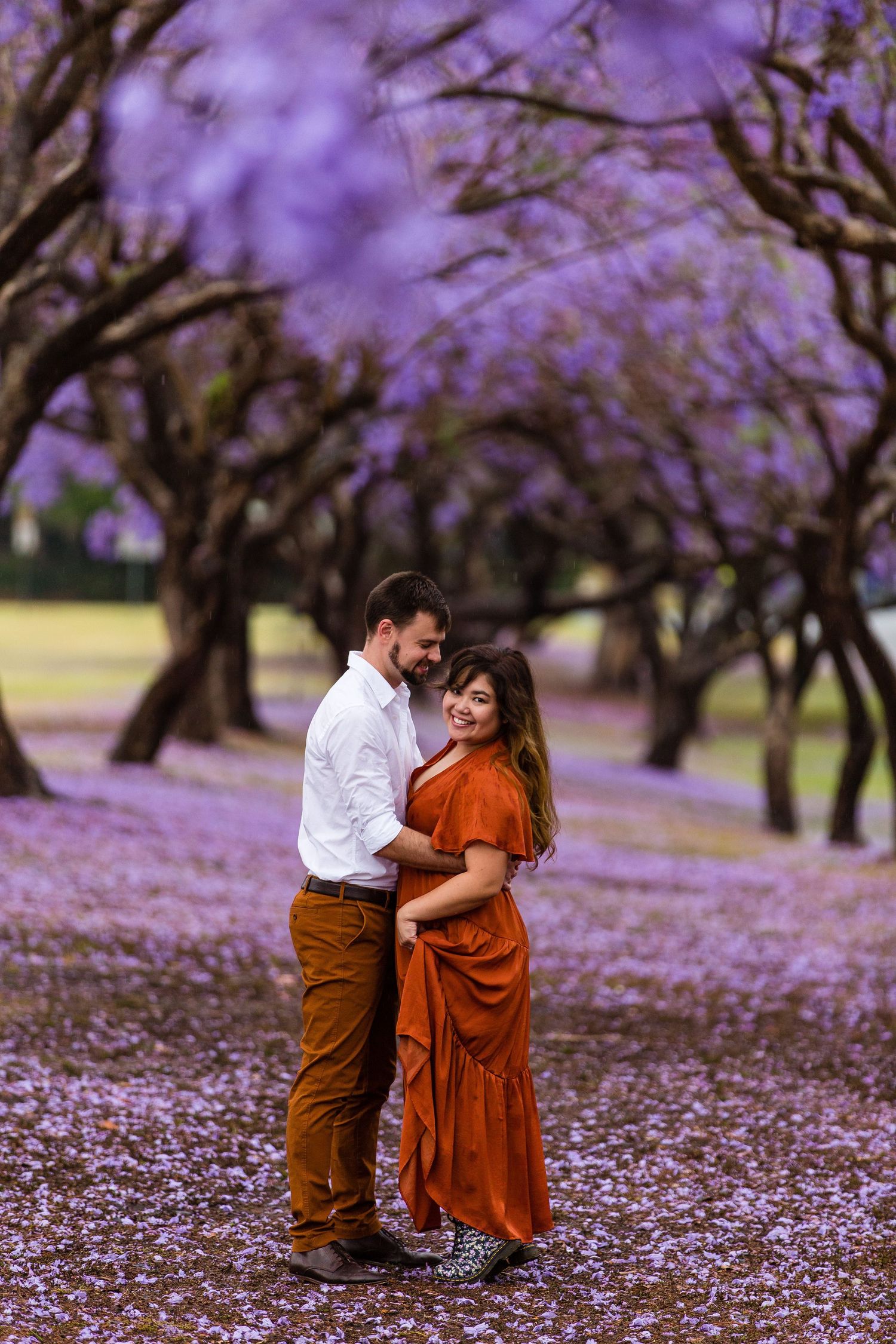 Romantic couple embracing under canopy of purple jacaranda blossoms in a dreamy garden setting.