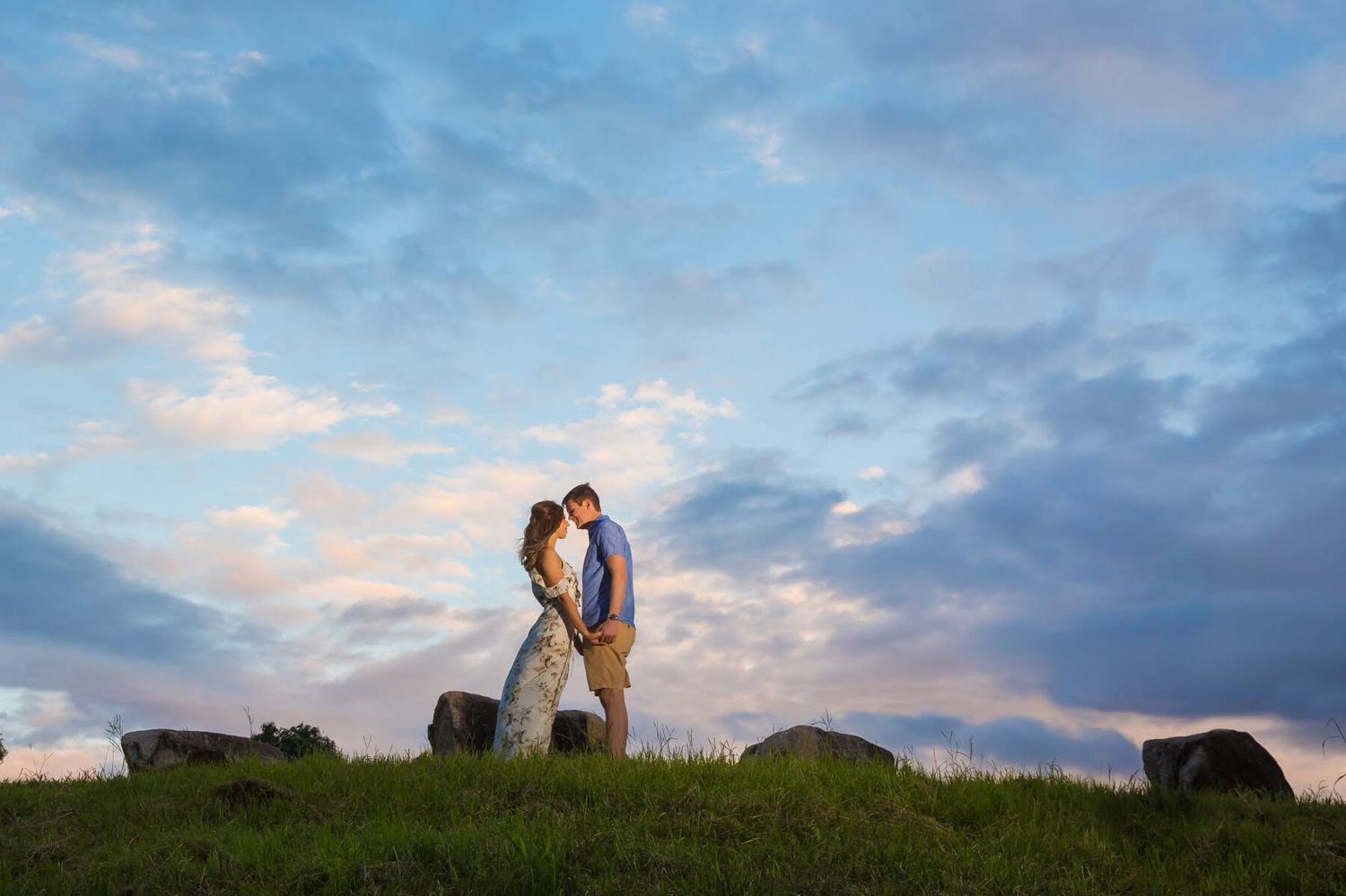 A romantic couple embraces on a grassy hilltop against a dramatic sunset sky with clouds.
