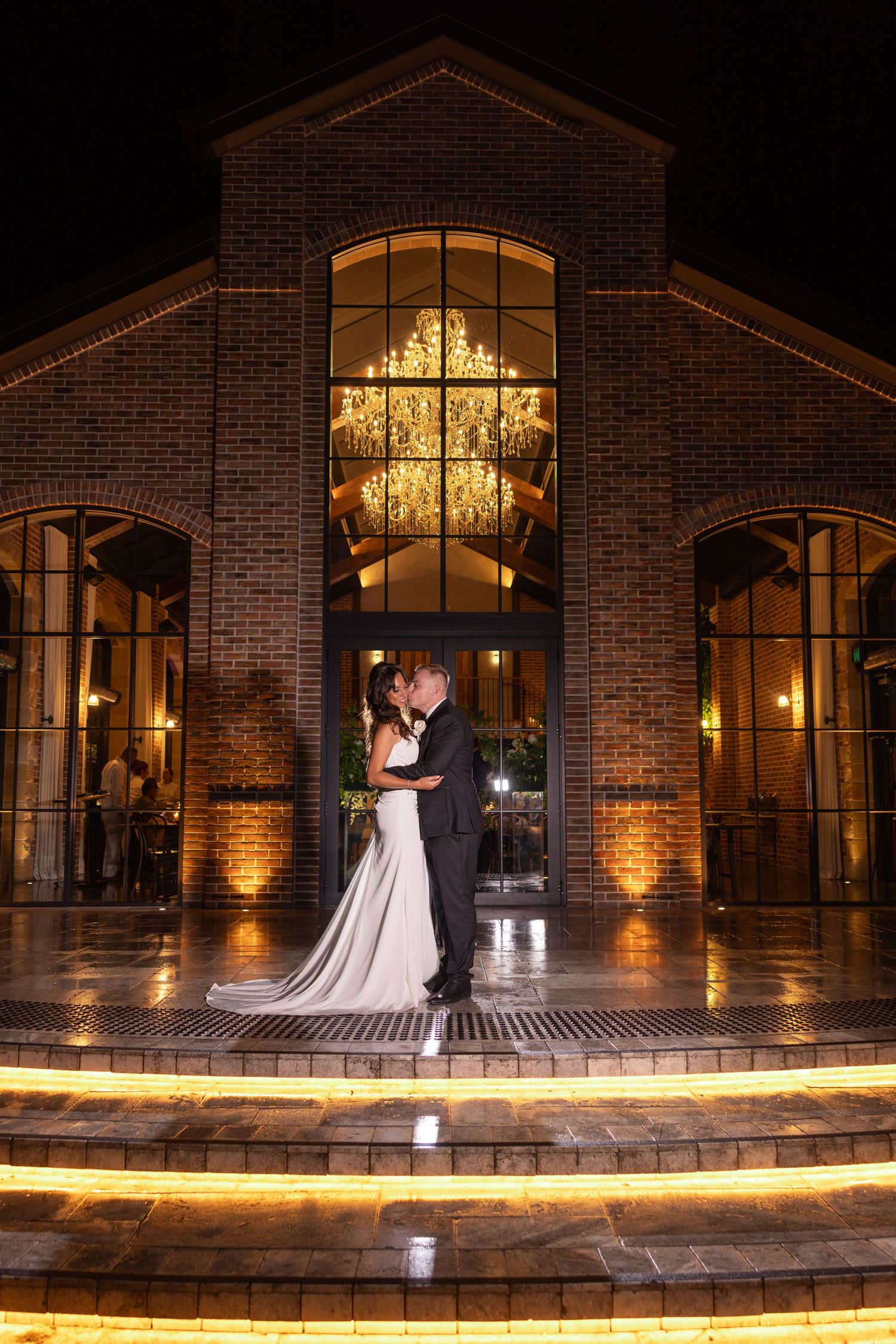A couple embraces at night outside a brick venue with illuminated windows and a grand chandelier visible through arched glass.