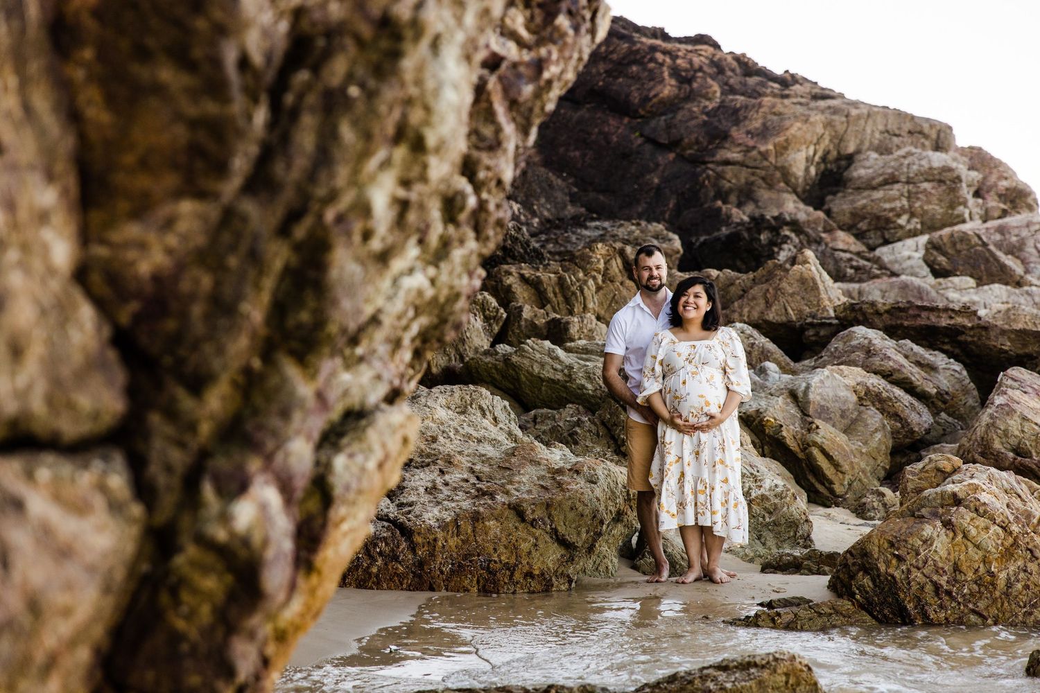 Person in a white dress stands among large coastal rock formations at the beach during golden hour.