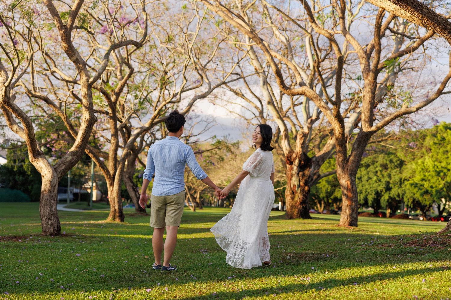 A couple walks hand in hand through a sun-dappled park with flowering trees on a warm afternoon.