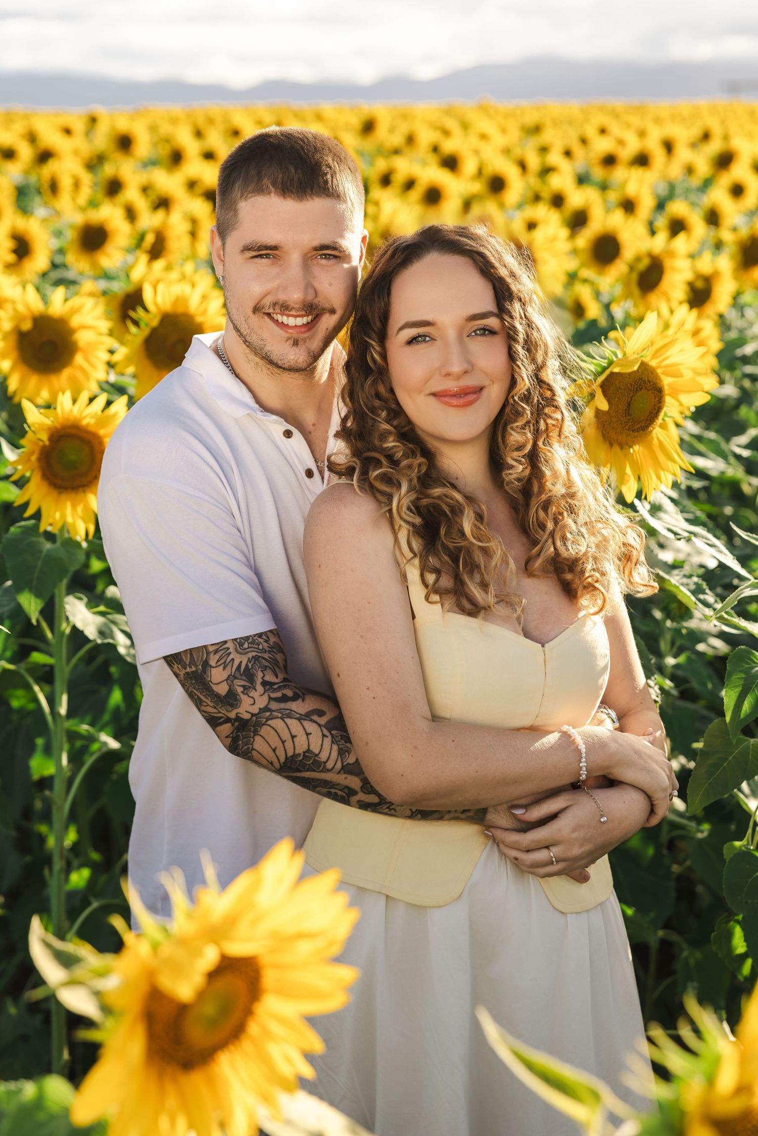 Happy couple embracing in a sunflower field, man with tattoo in white shirt and woman in yellow dress.