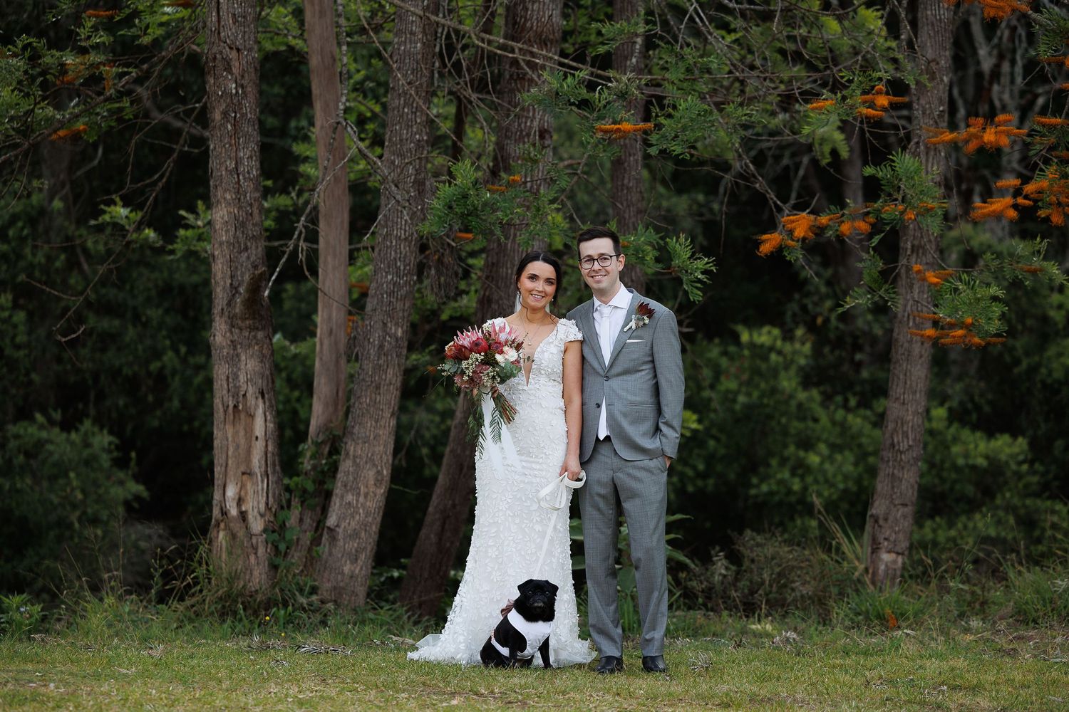 Bride and groom posing with black dog in white wedding dress and gray suit before lush forest backdrop.