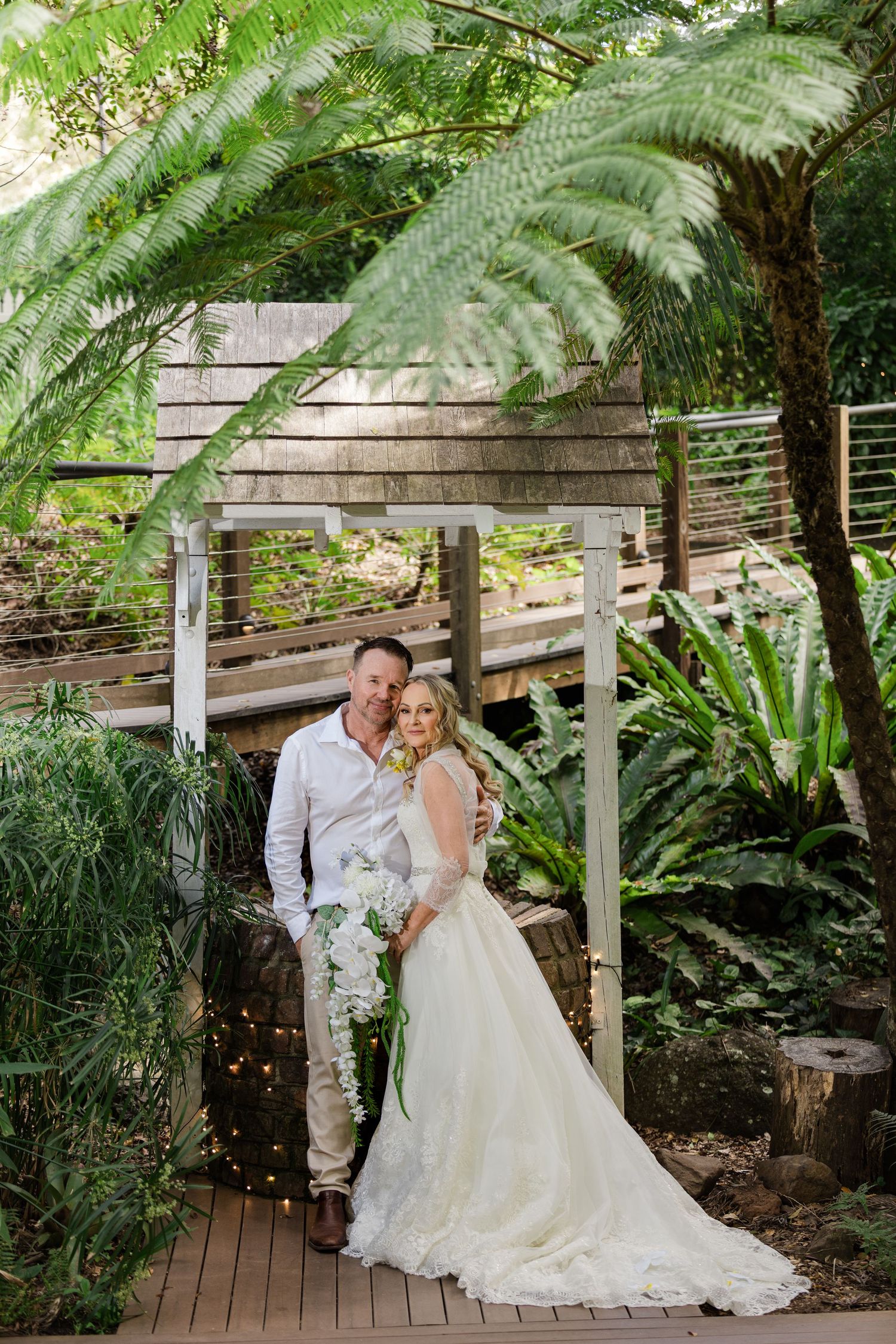 Bride and groom pose together in a lush garden wedding venue with tropical ferns and a wooden bridge backdrop.