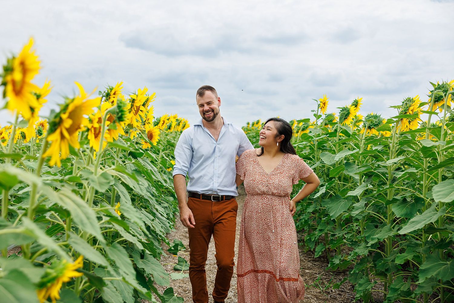 Two people walk together through a vibrant field of tall sunflowers on a cloudy summer day.