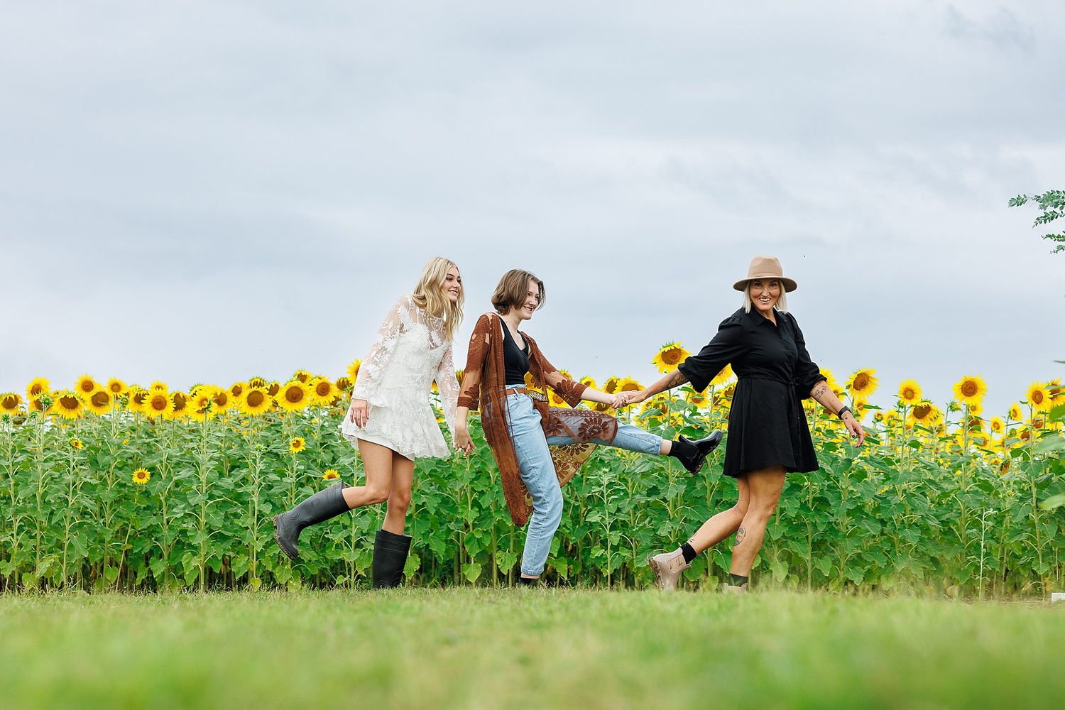 Three people dance together holding hands along the edge of a vibrant sunflower field on a cloudy summer day.