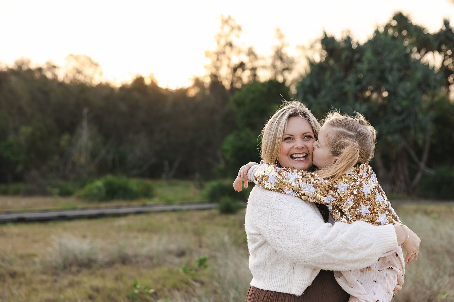 A parent and child share a joyful embrace in a field at sunset, both wearing cozy sweaters.