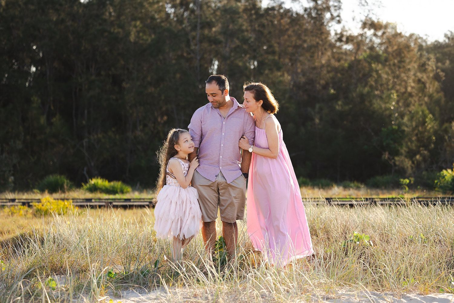 Three people in pink and purple outfits stand together in a sunlit grassy meadow during golden hour.