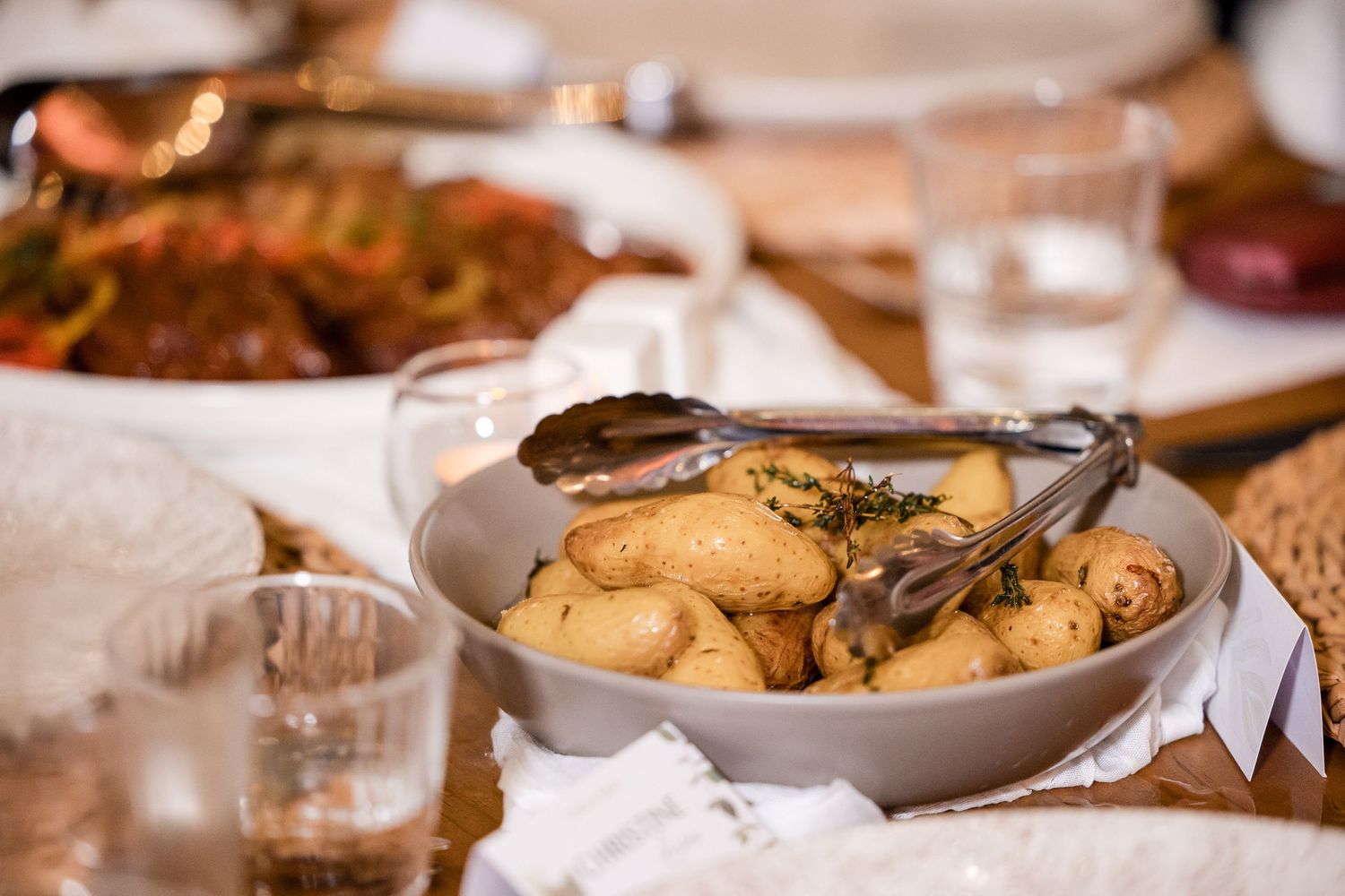 A bowl of roasted baby potatoes with herbs and tongs on a festive dining table with glasses and dishes.