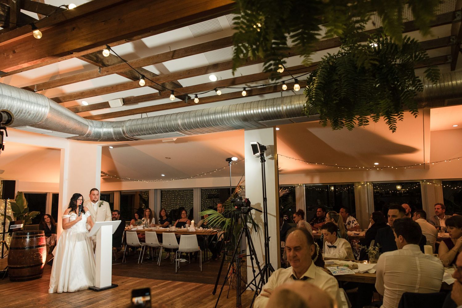 Two brides in white gowns give a speech at an elegant wedding reception with warm lighting and lush greenery.
