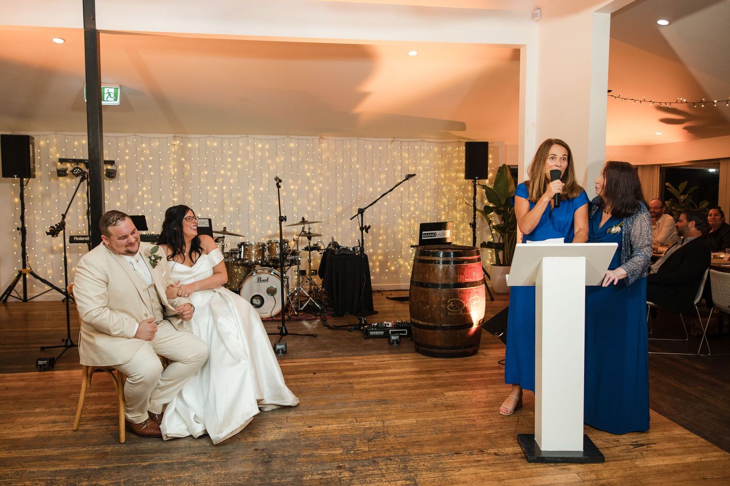 Wedding reception speech by bridesmaid in blue dress while bride and groom sit together in elegant venue.