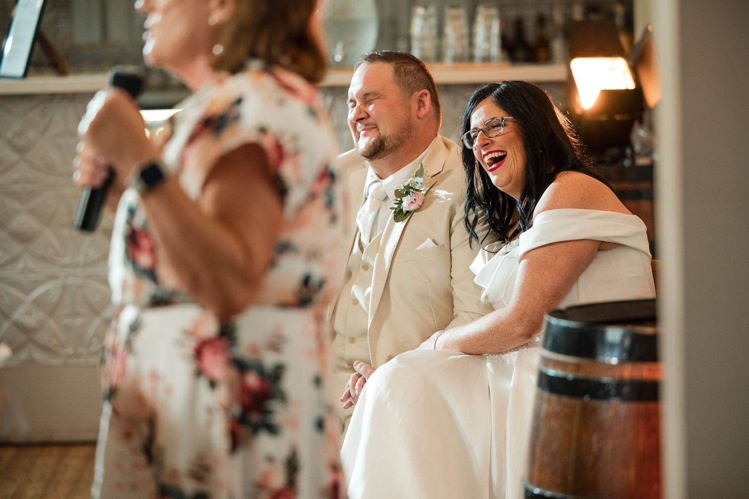 Bride and groom laughing joyfully during wedding reception speech at elegant venue.