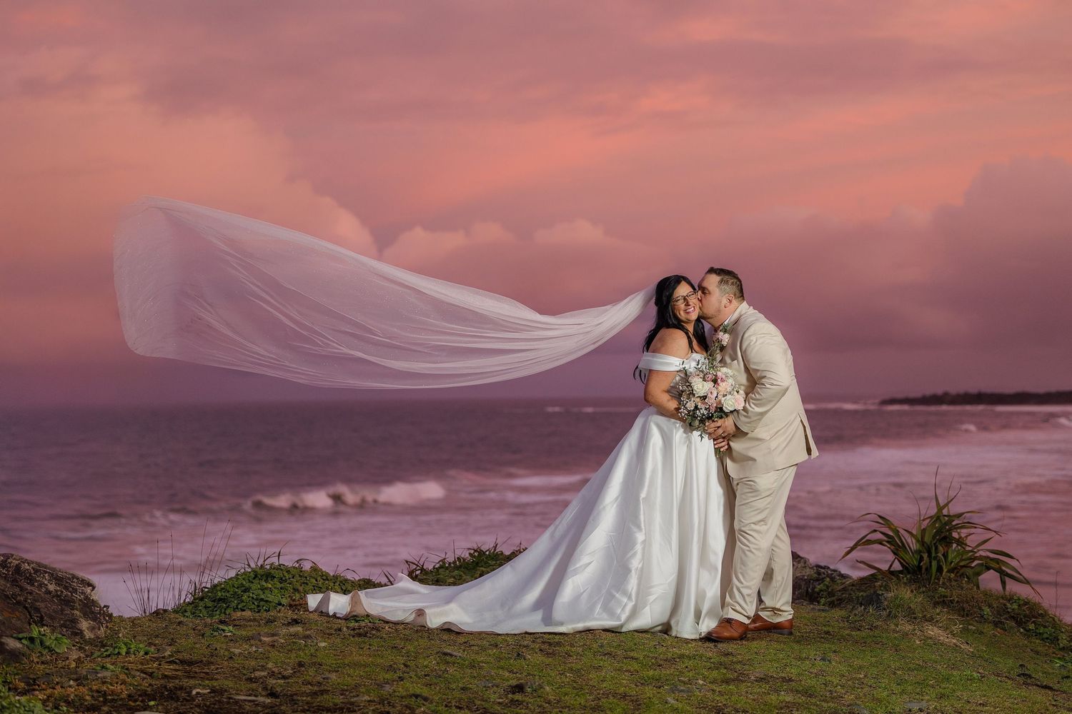 Bride and groom share a romantic kiss at sunset on a coastal cliff, with her veil flowing dramatically in the wind.
