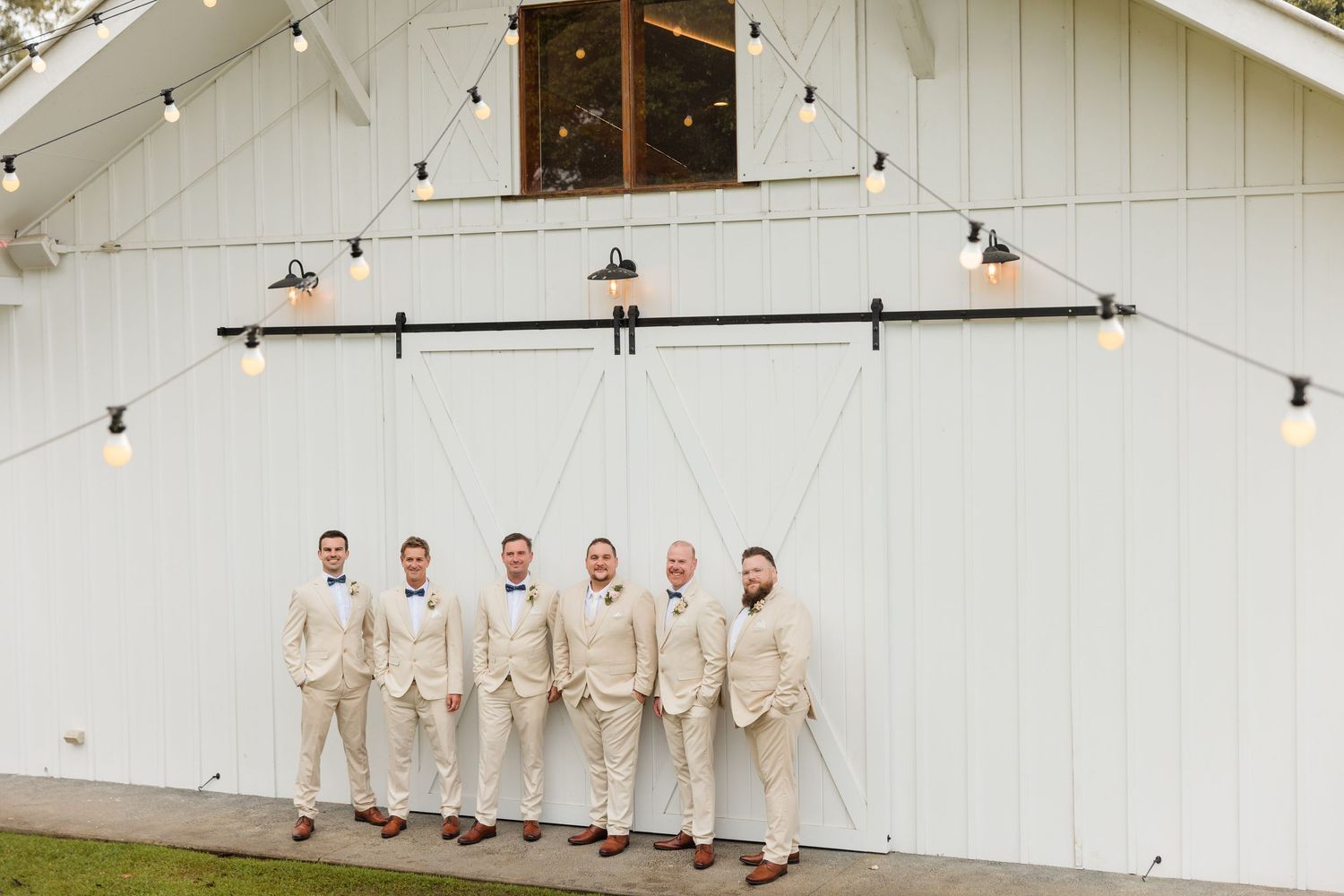 Groom and groomsmen in matching beige suits with bow ties posing in front of a white barn wedding venue.