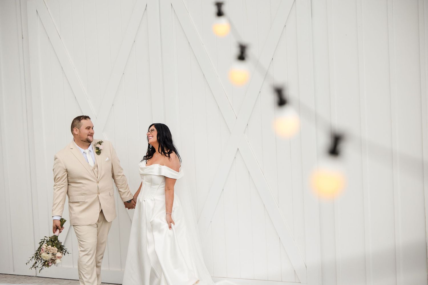 Bride and groom holding hands smiling at each other in front of white barn doors with string lights.