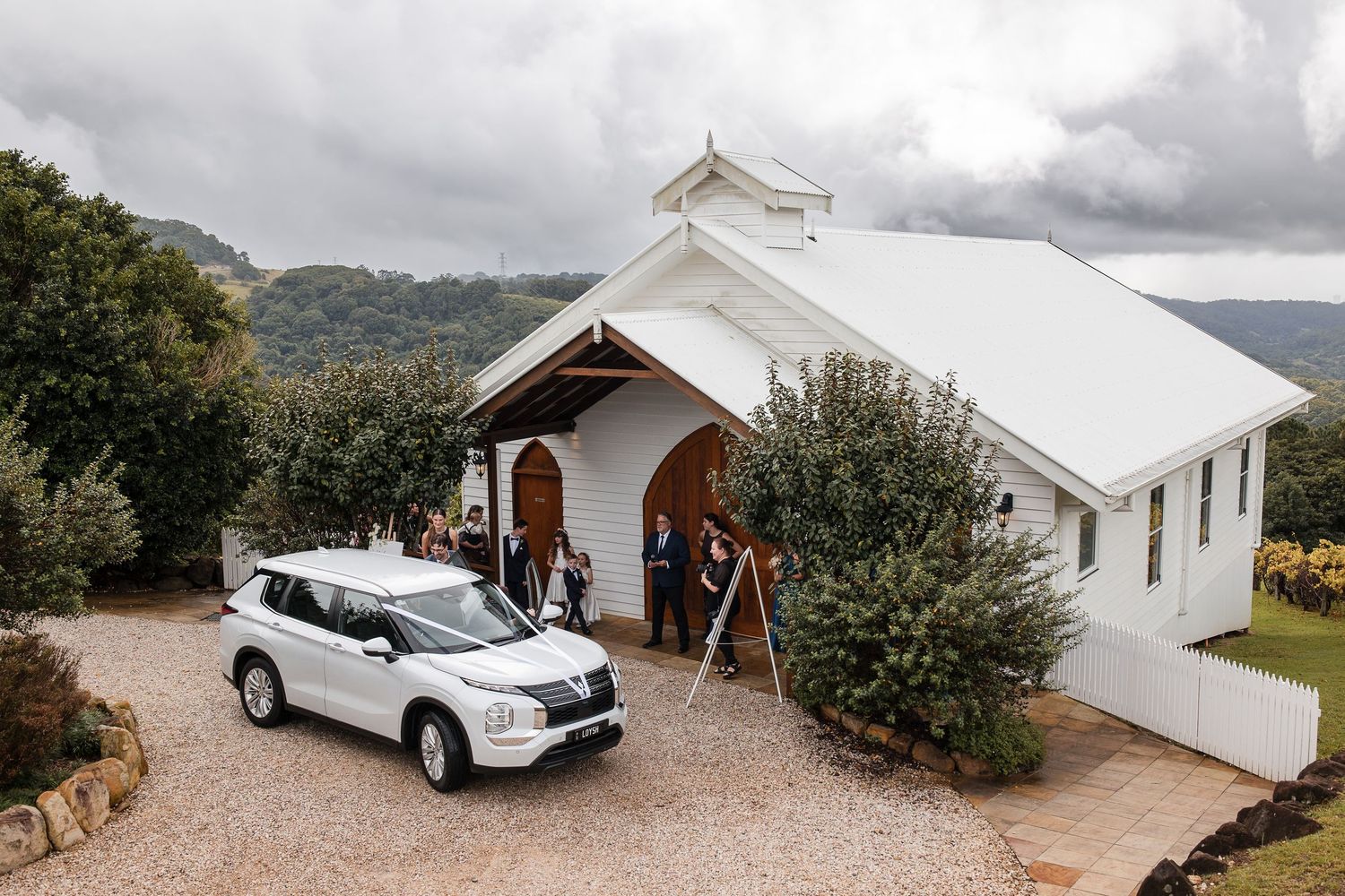 White Mitsubishi SUV parked outside a charming white chapel with wedding guests gathered at the entrance.