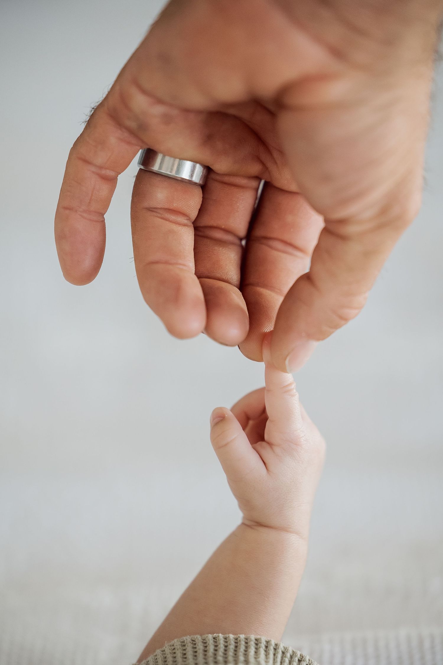 A baby's tiny hand gently gripping the pinky finger of an adult hand wearing a silver ring.