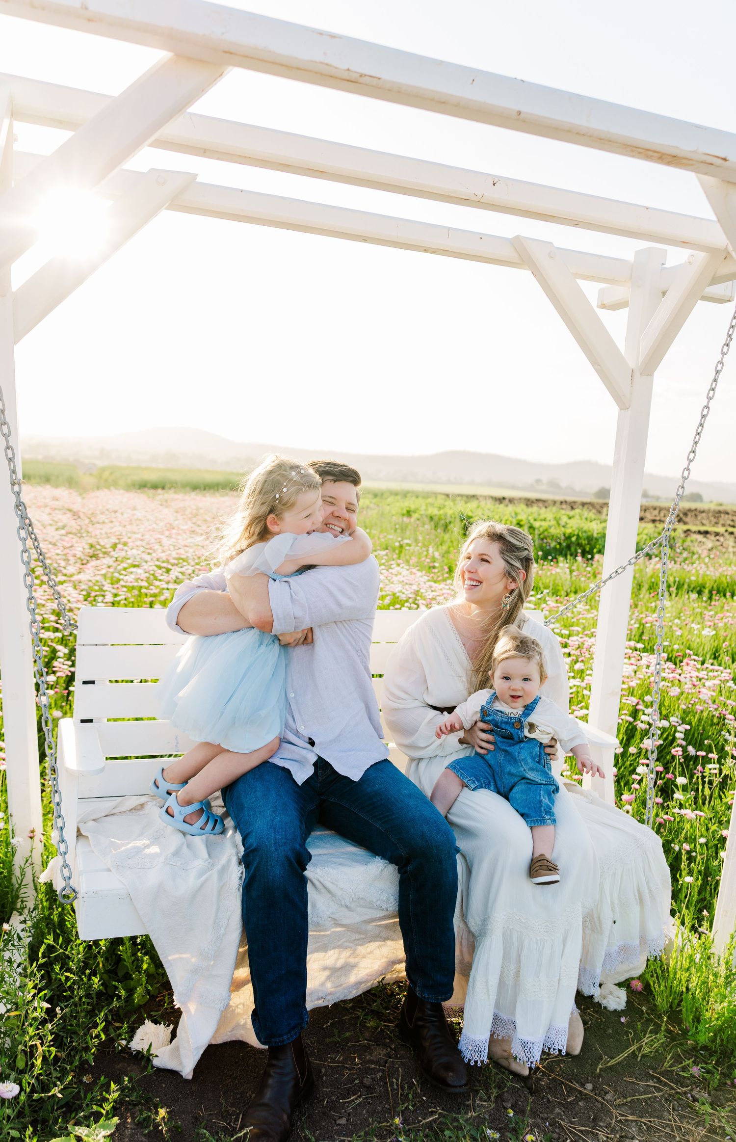 Family of four sitting on a white garden swing surrounded by wildflowers on a sunny day.