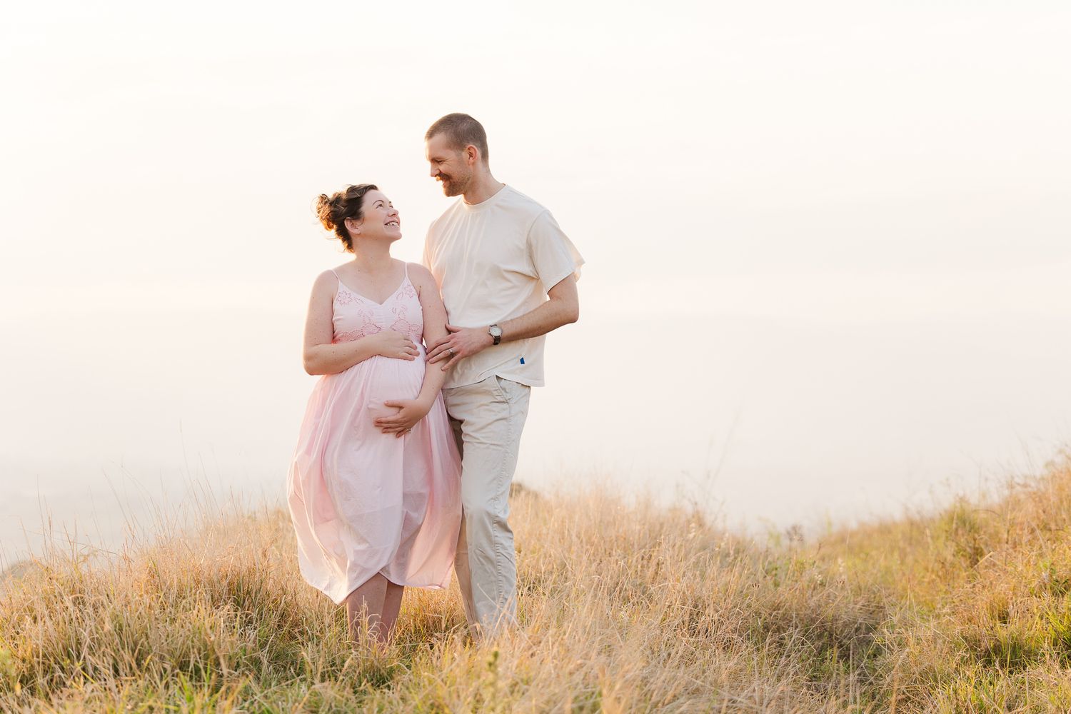 A couple stands together in a golden field during sunset, sharing a tender moment in warm lighting.