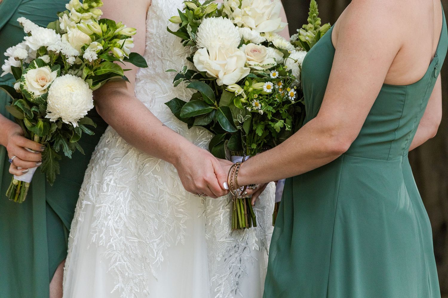 Bride in lace gown with bridesmaids in green dresses holding white peony bouquets at an outdoor wedding.