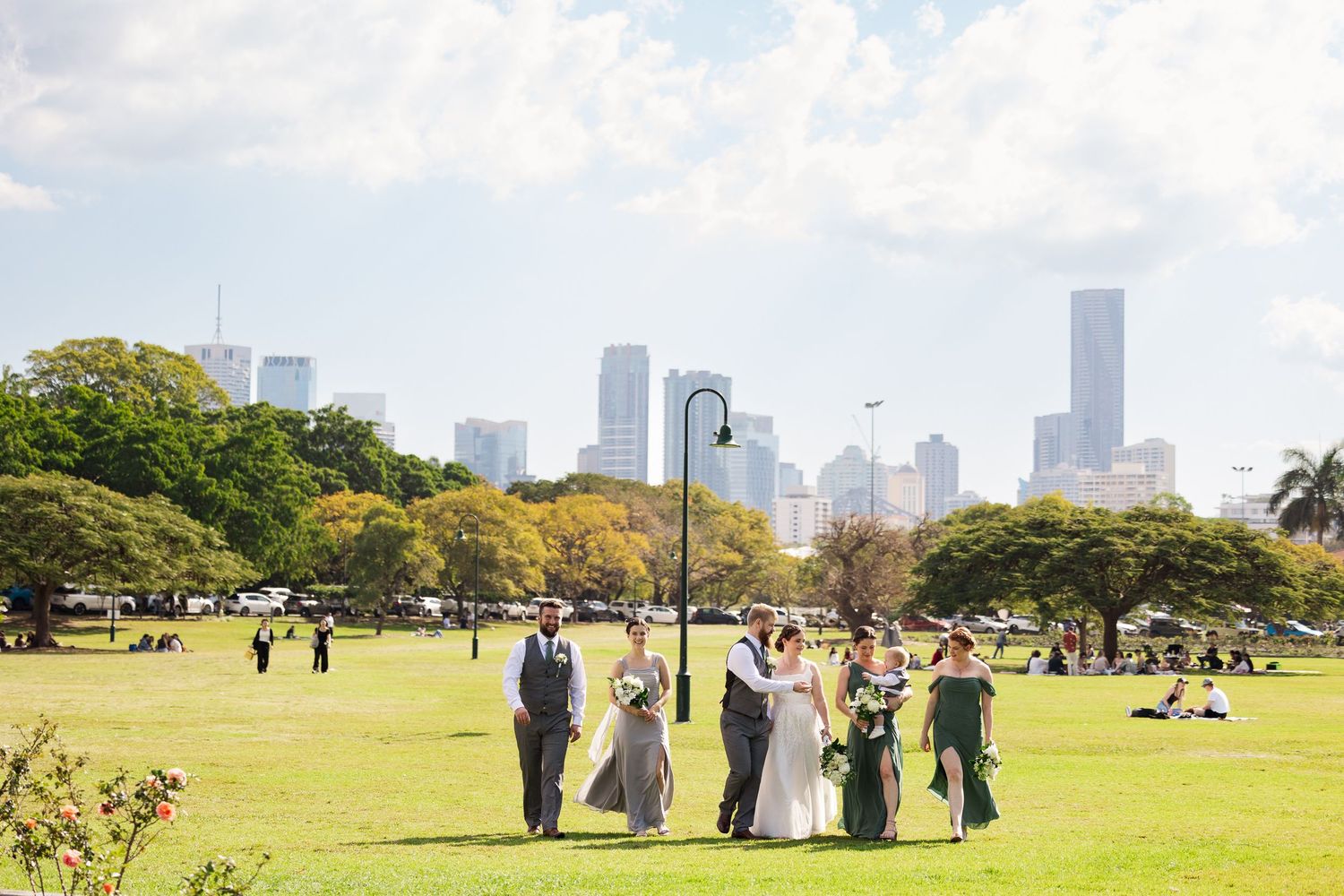 Wedding party poses on green lawn with city skyline backdrop on a sunny day.