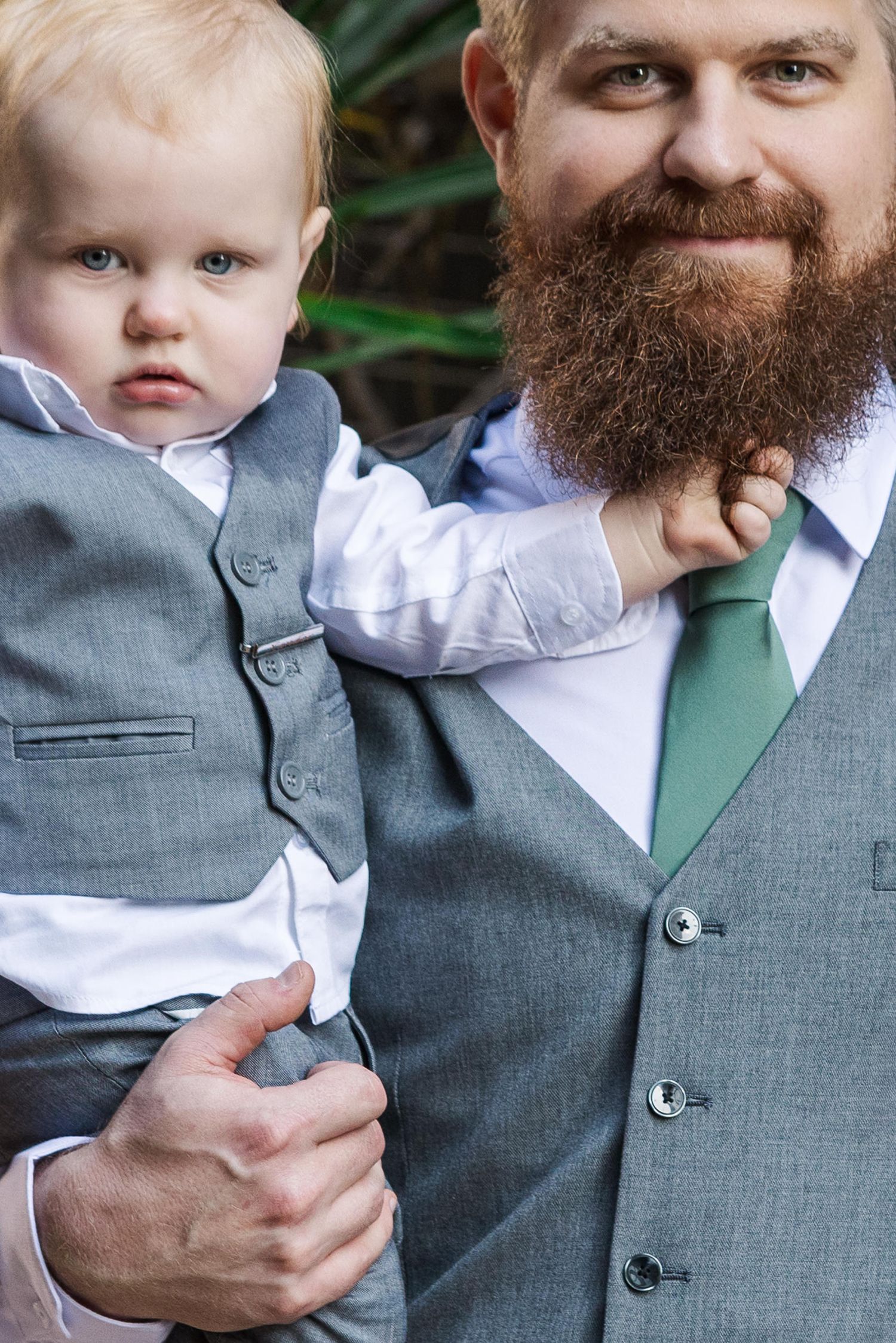 Bearded man in gray suit and green tie holds baby boy in matching gray vest suit at formal event.