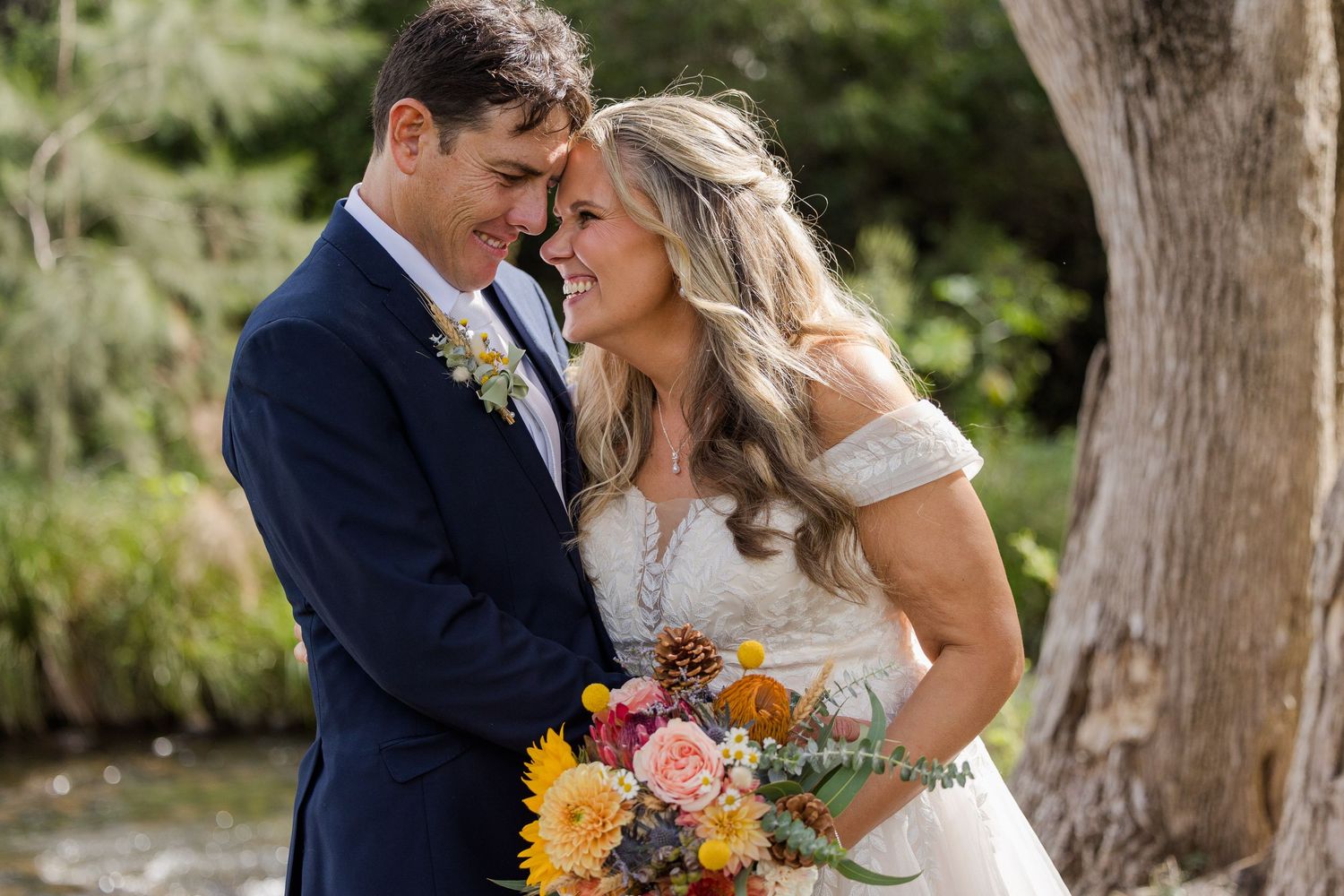 Bride and groom smiling intimately outdoors, bride holding vibrant fall bouquet with sunflowers, groom in navy suit.