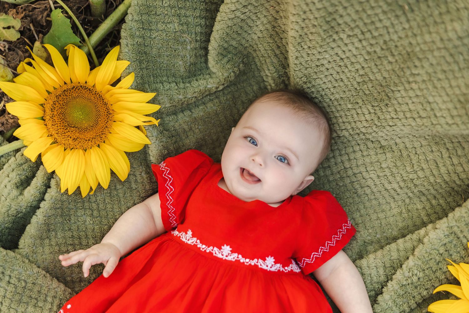 Adorable baby in red dress lying on green blanket surrounded by bright yellow sunflowers.