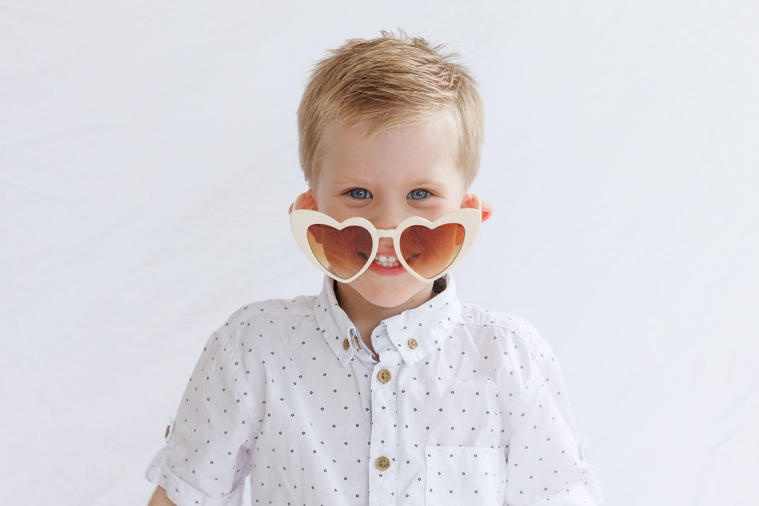 A young child wearing a white polka dot shirt and heart-shaped pink sunglasses poses against a white background.