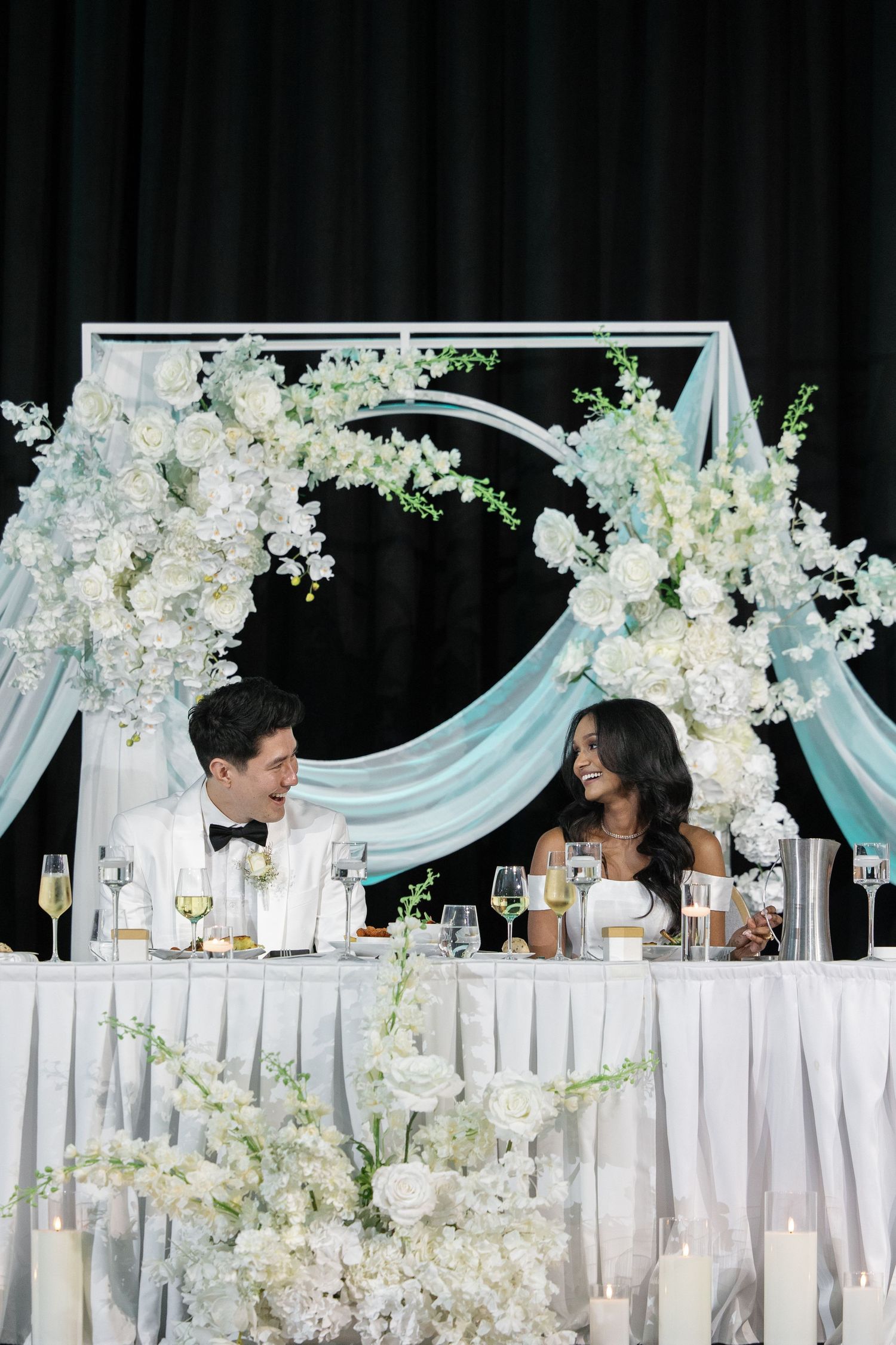 Elegant wedding reception setup with white floral arrangements and light blue draping decorating a long head table.