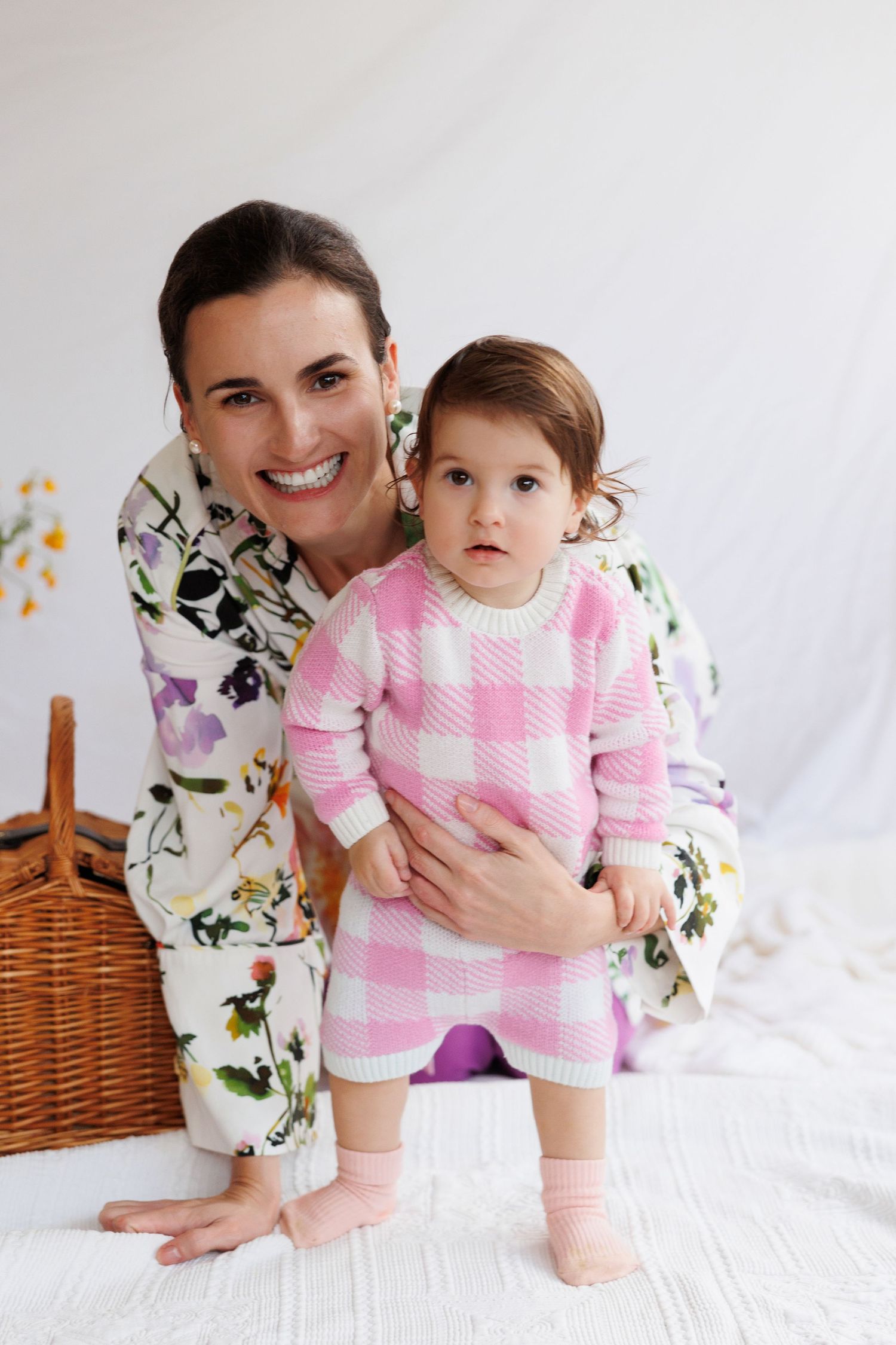 A smiling woman in floral dress kneels beside a toddler in pink gingham knit romper on a white backdrop.