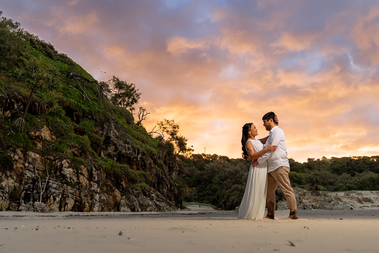 A couple embraces on a beach at sunset with dramatic cliffs and orange-pink sky in the background.