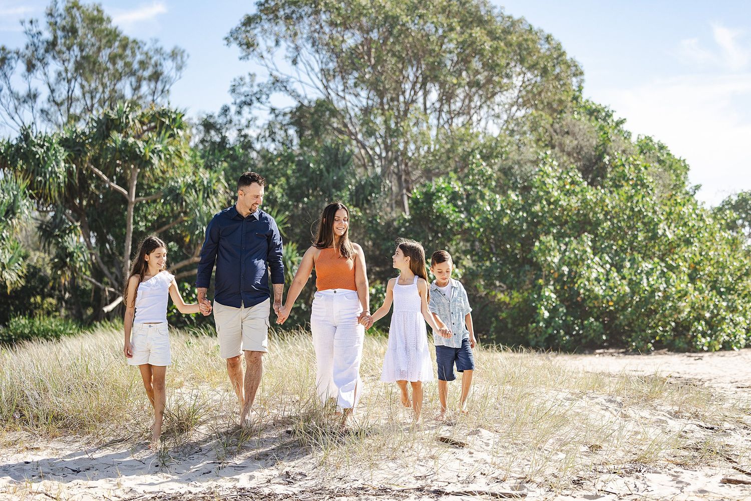 Family walking together on sandy beach path surrounded by lush green trees and vegetation on sunny day.