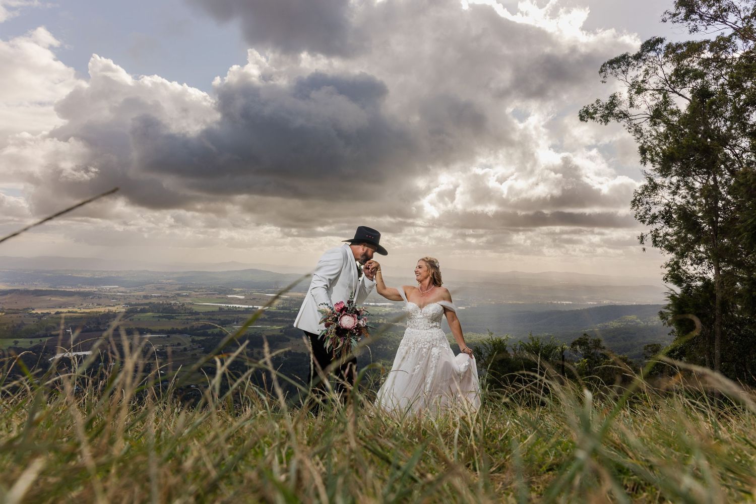 Bride and groom dancing on a hilltop with dramatic cloudy skies and scenic valley views in the background.
