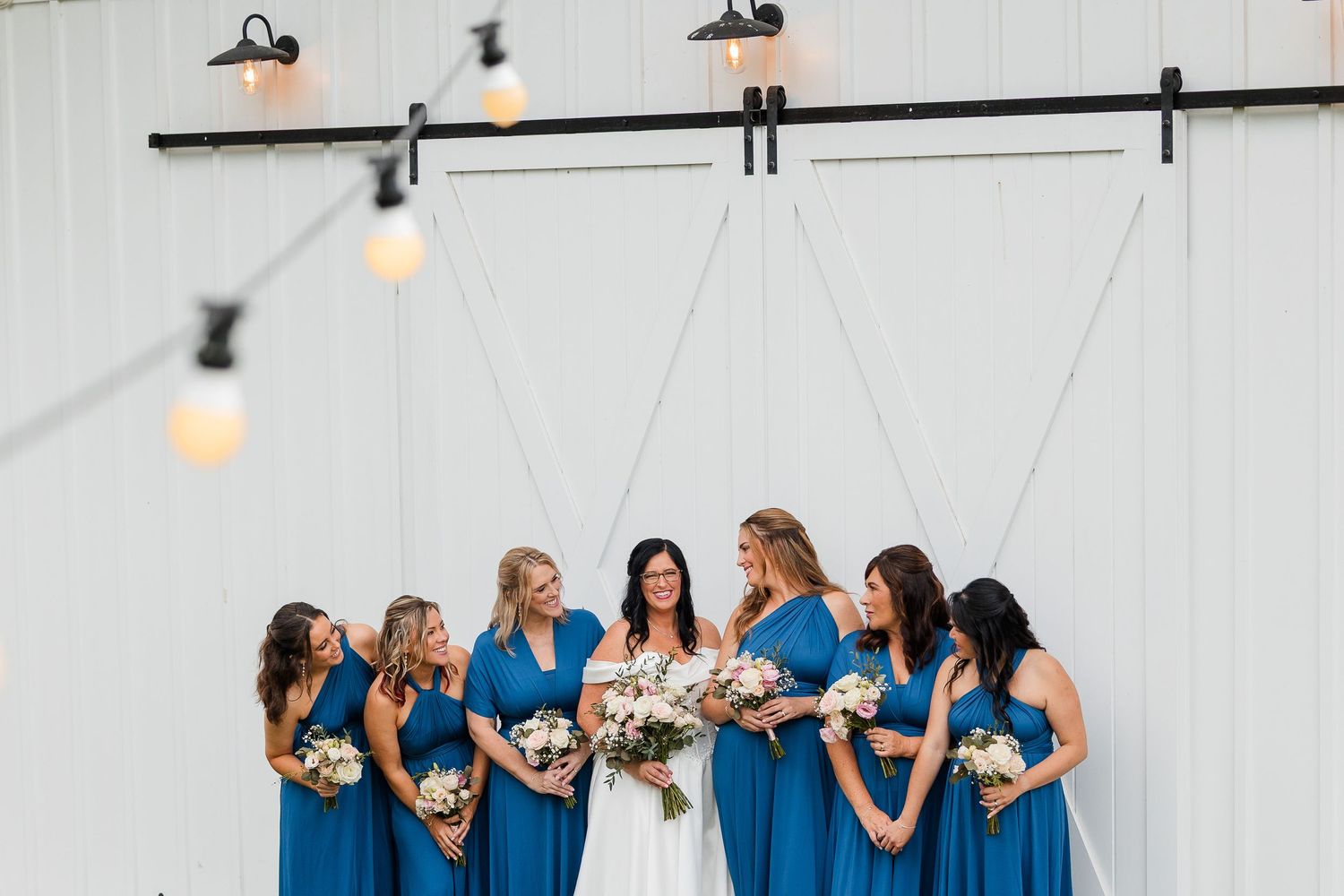Bride in white gown with bridesmaids in blue dresses holding bouquets outside a white barn wedding venue.
