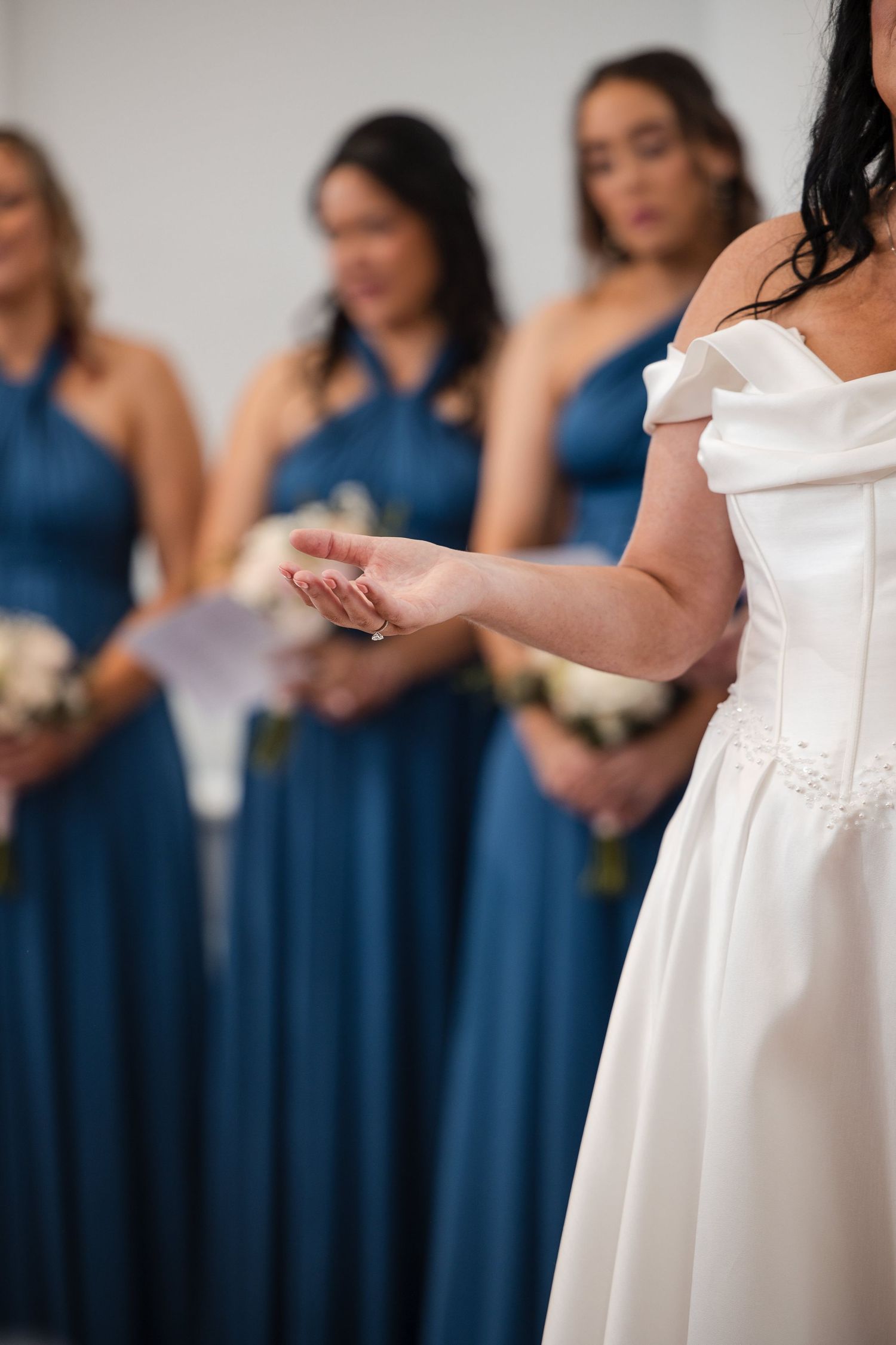 Bride in white off-shoulder gown gestures to bridesmaids in blue dresses holding bouquets at wedding.