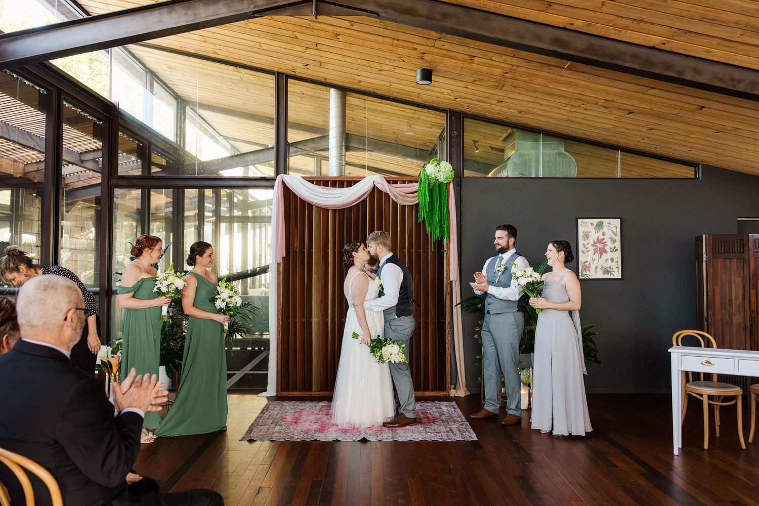 Wedding ceremony with couple kissing at floral arch, bridesmaids in green dresses, officiant, and guests in rustic venue.
