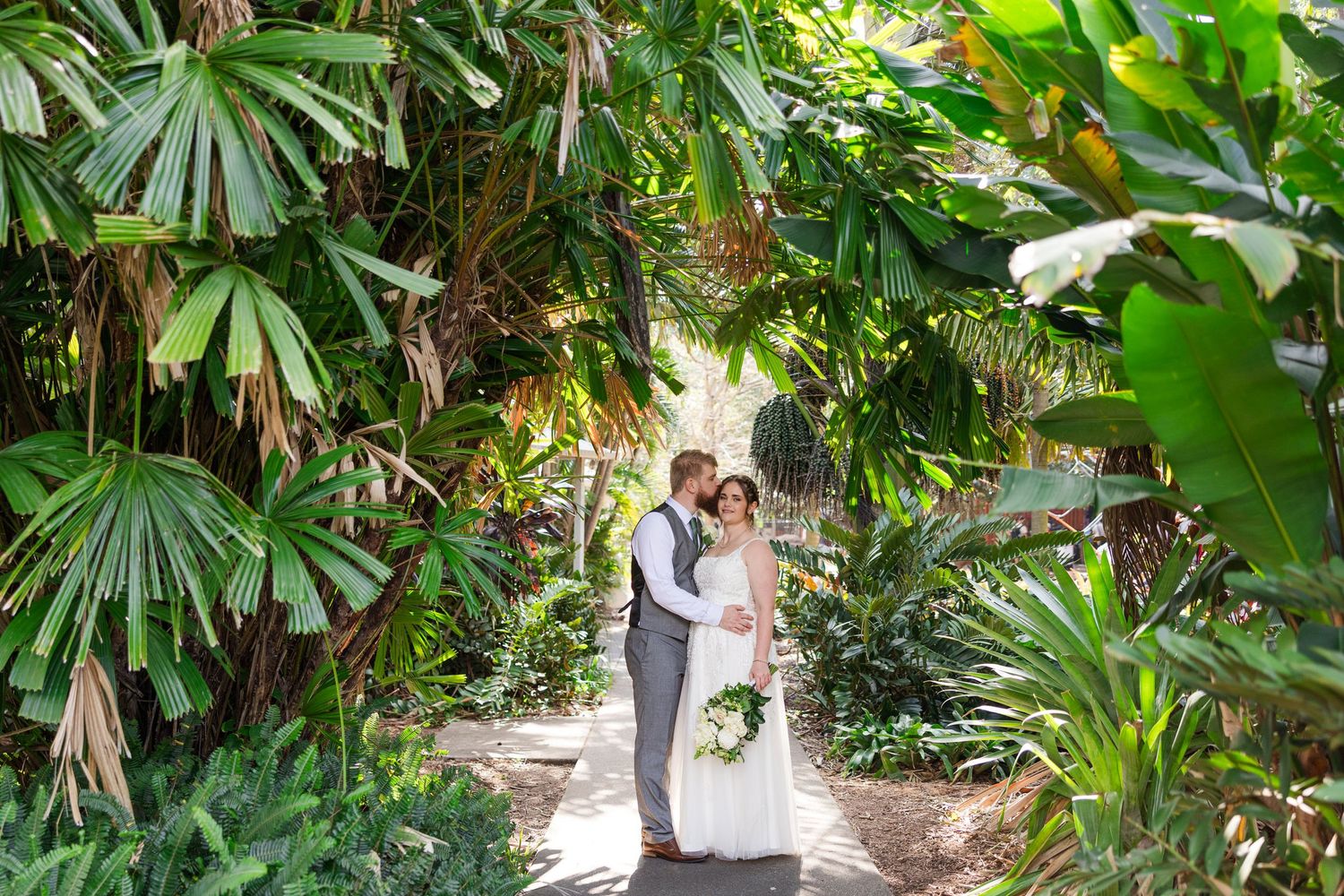 Bride and groom share a romantic moment surrounded by lush tropical greenery on their wedding day.